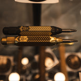Two black and polished brass auto centre punches, displayed horizontally on a reflective surface with a blurred indoor background.