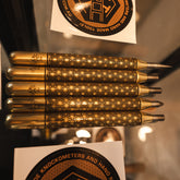 Five brass punches and scribe with ornate Kumiko-patterned engravings are displayed horizontally on a glass surface, with circular brand stickers visible in the background.