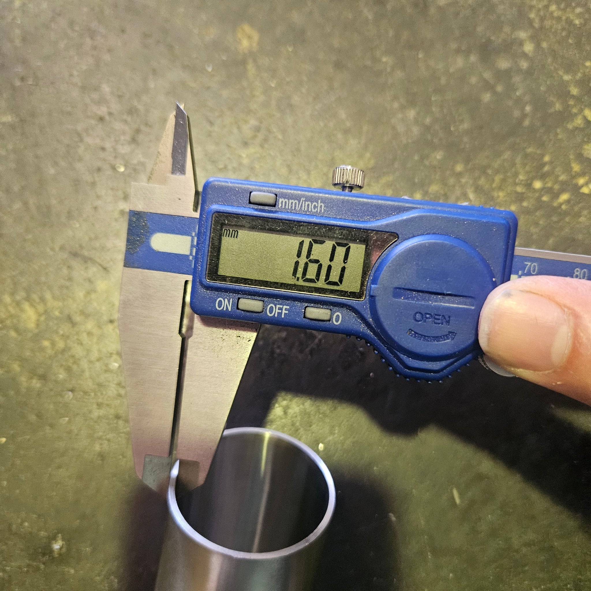 A blue digital caliper held by a hand, measuring the wall thinkness of a metal tube. The caliper display shows a measurement of 1.6mm