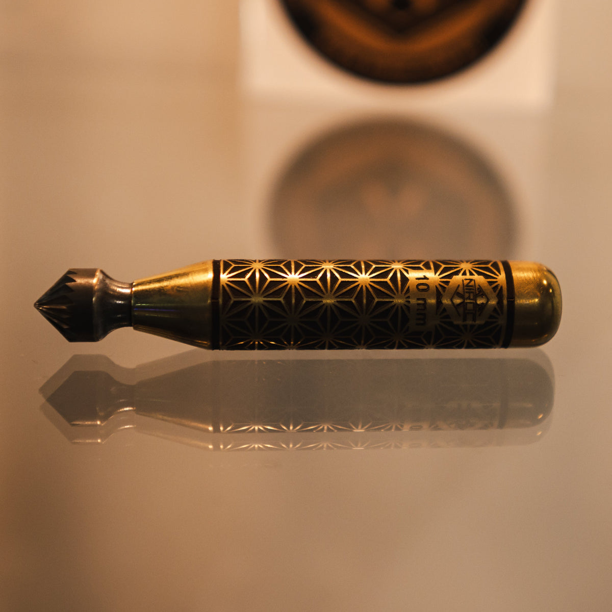 A brass and black decorative tool with intricate starburst engravings, resting on a reflective surface with a blurred circular emblem in the background.