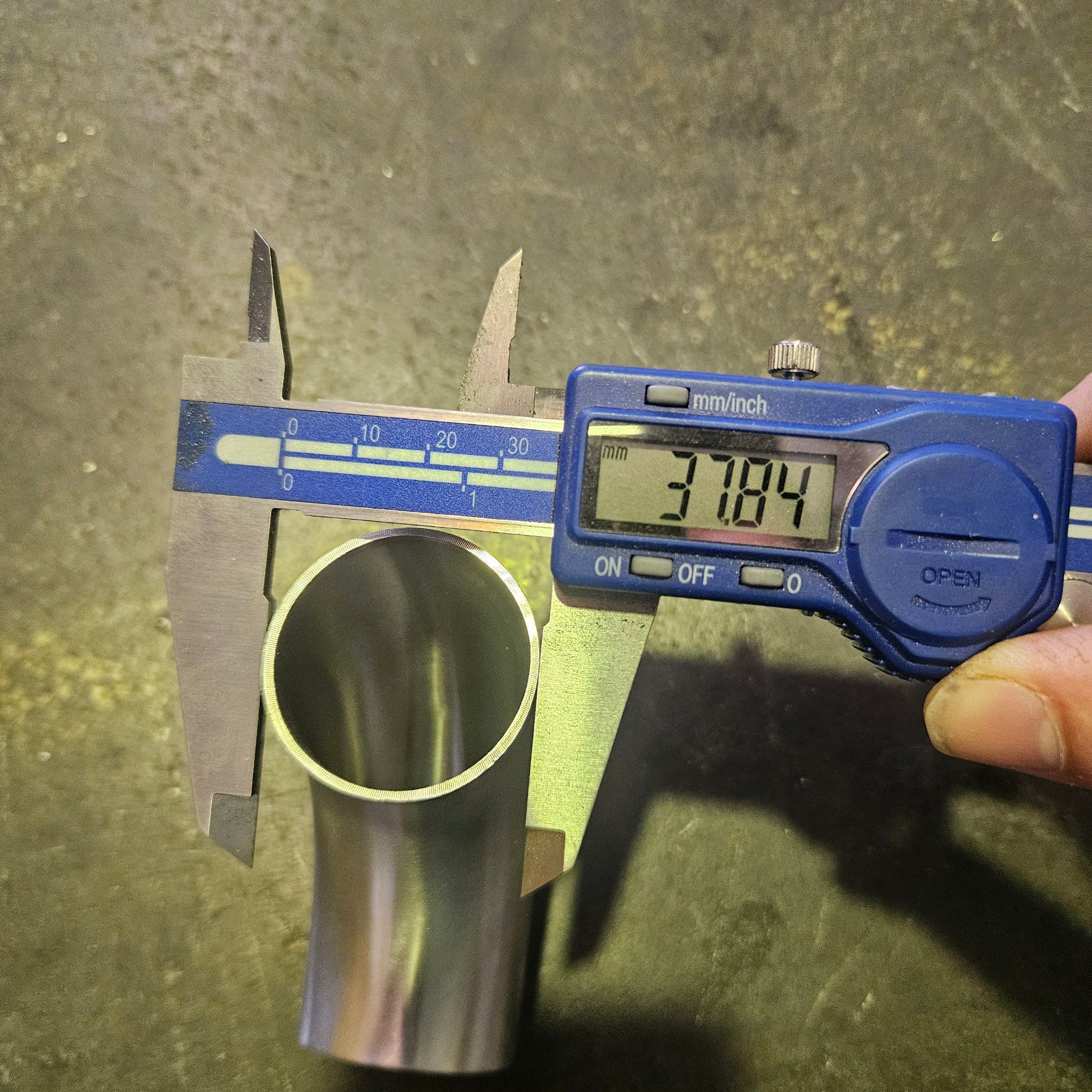A stainless steel pipe or tube being measured with a blue digital caliper. The caliper display reads 38.1mm.