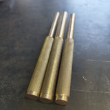 Three brass drifts with knurled textured grips and smooth metal shafts, laying down on a dark steel workbench.