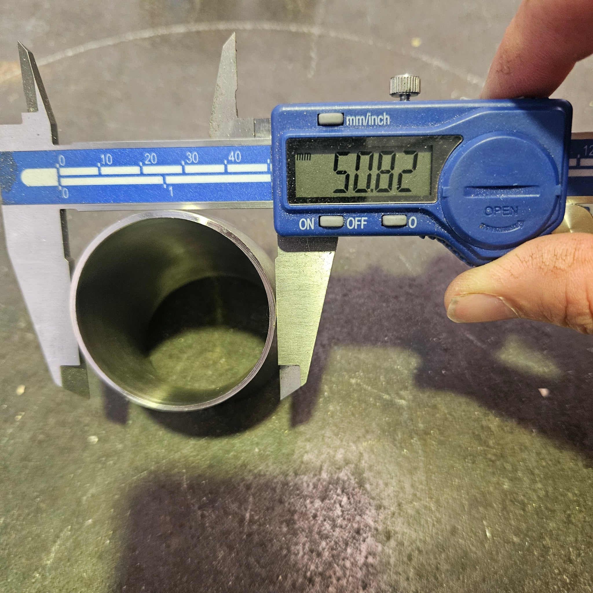 A digital caliper measuring the outer diameter of a metal tube, with a hand holding the calliper. The caliper has a blue display screen reading 50.8mm and silver metal jaws, and the tube is positioned on a dark, textured surface.