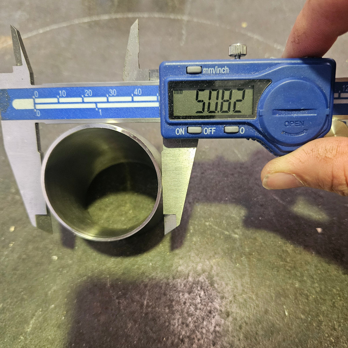 A digital caliper measuring the outer diameter of a metal tube, with a hand holding the calliper. The caliper has a blue display screen reading 50.8mm and silver metal jaws, and the tube is positioned on a dark, textured surface.