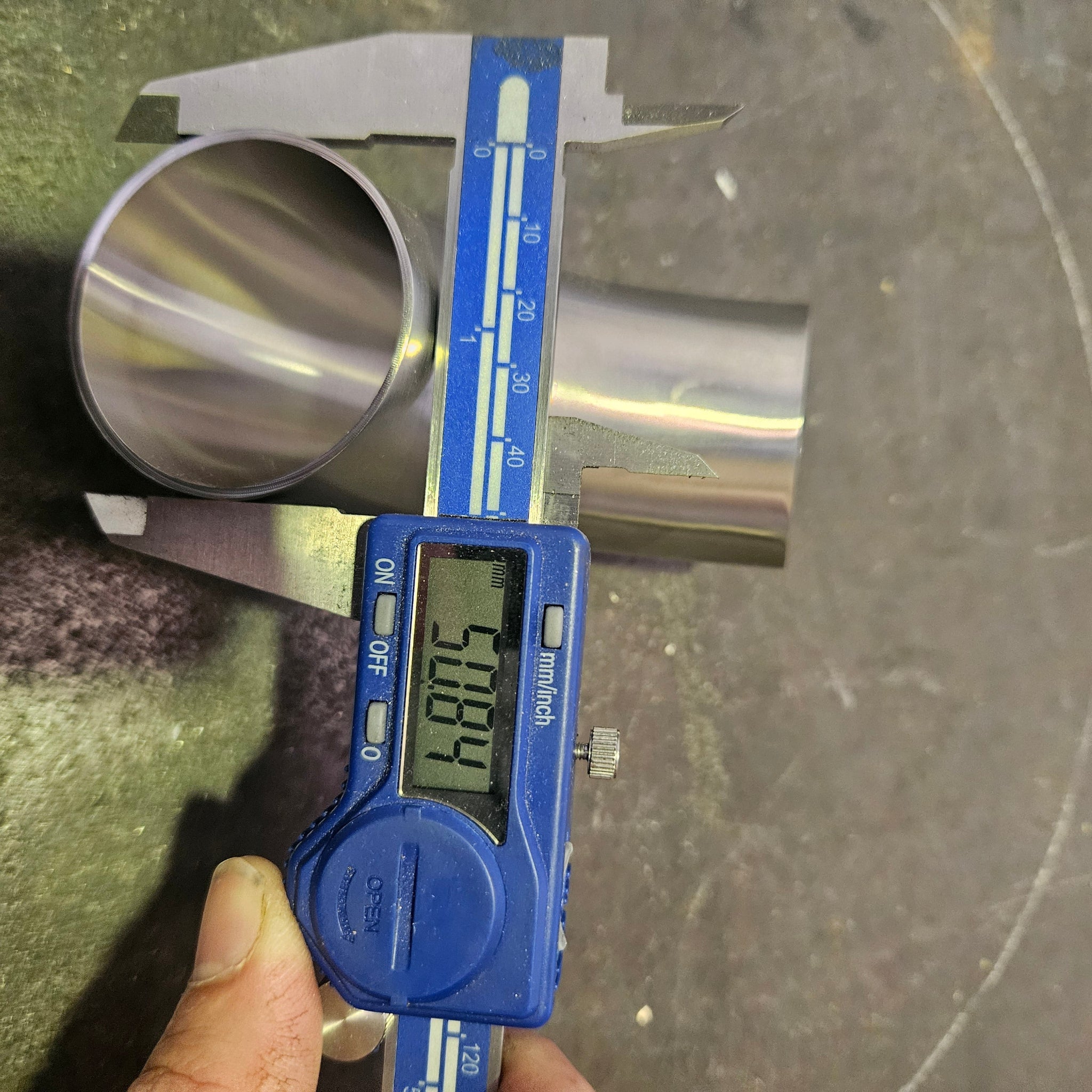 A blue digital caliper is measuring the diameter of a shiny metal tube, with a hand holding the caliper at the bottom left. The caliper display shows a measurement of 50.8mm.