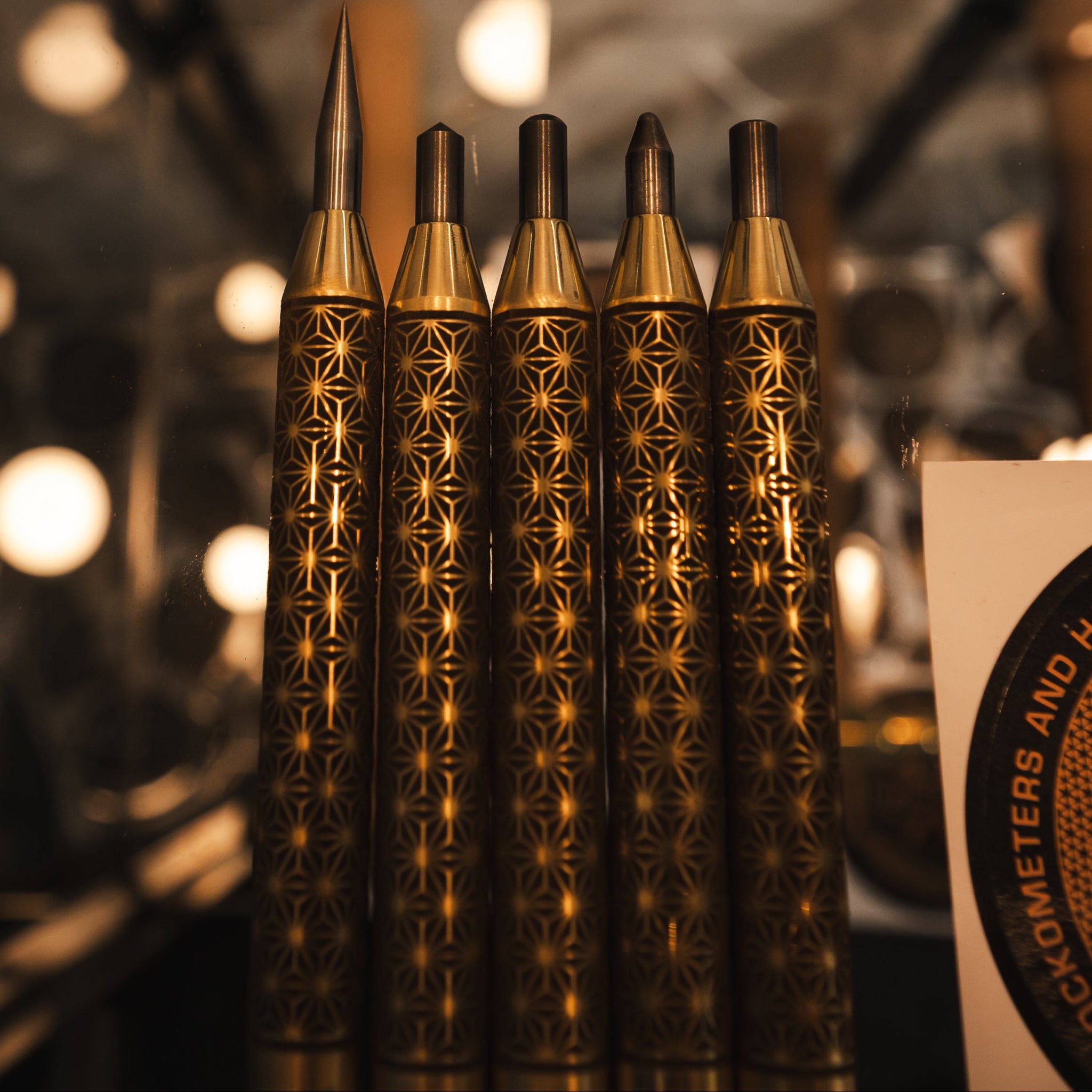 Five decorative blackened and polished brass precision scribe and punch set with intricate geometric patterns on their handles, arranged upright in a row against a warm, blurred background.