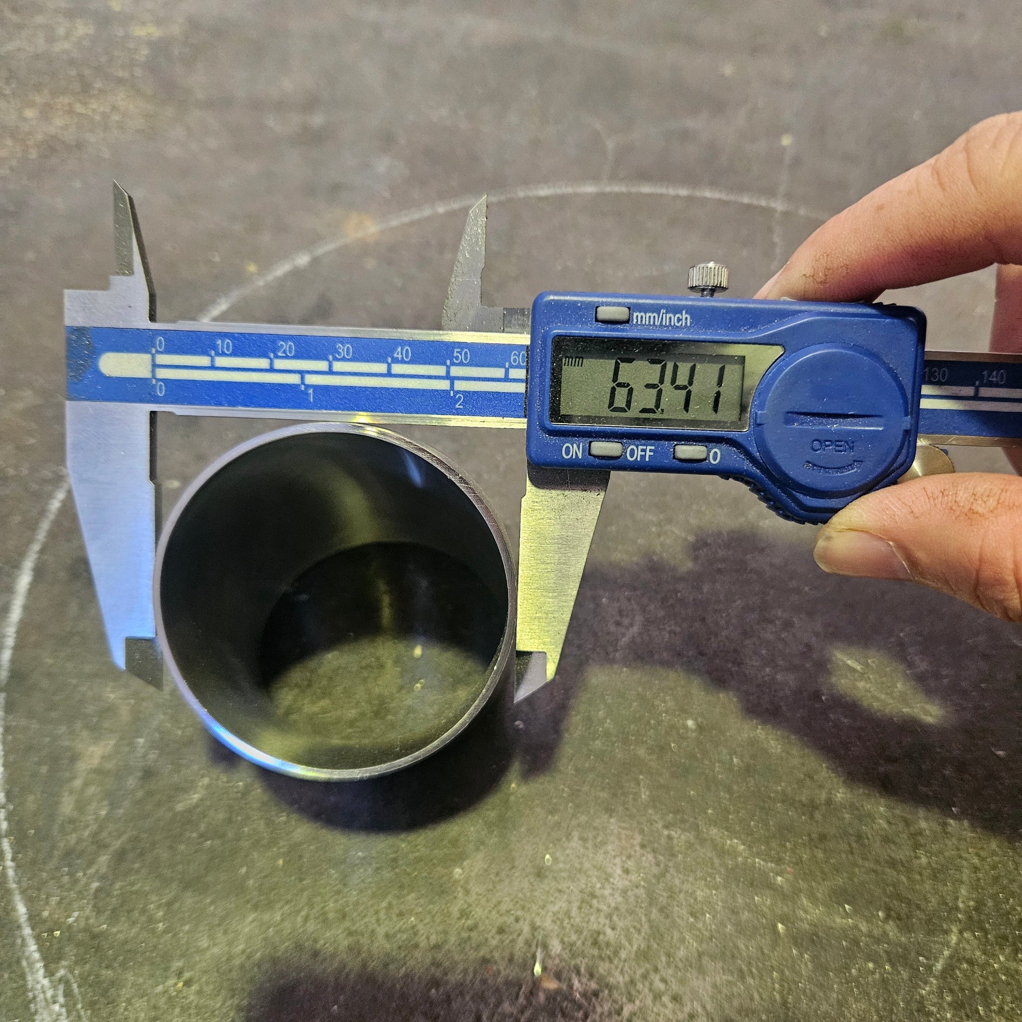 A hand holding a blue digital caliper measuring the diameter of a stainless steel 45 degree bend tube, with the screen displaying 63.5 mm.
