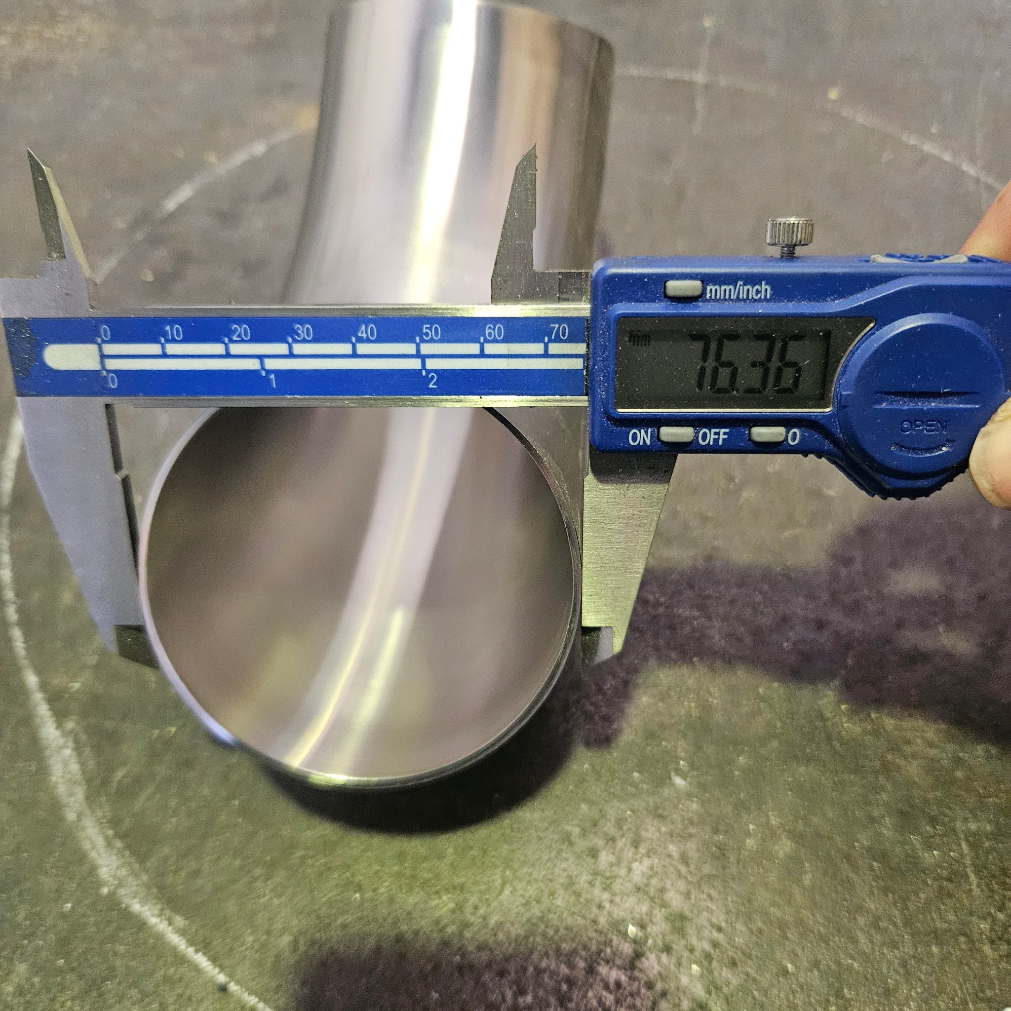A digital caliper measuring the outer diameter that reads 76.2mm of a polished stainless steel pipe, with a hand holding the caliper on a dark textured floor.