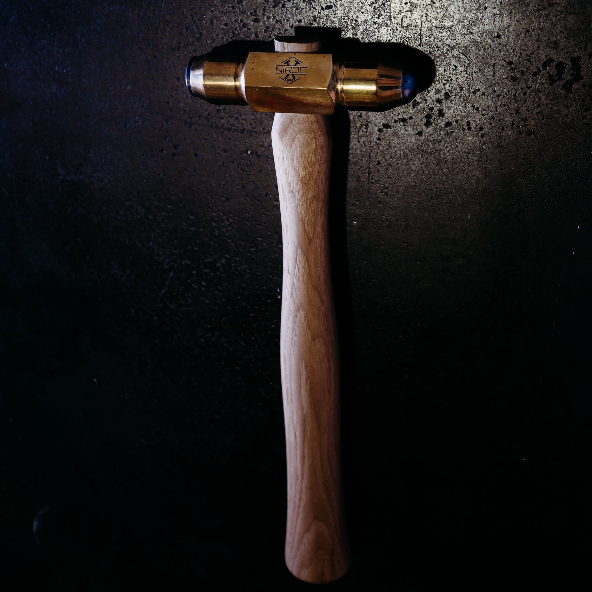 A wooden-handled hammer with a brass head and carbide faces, photographed against a dark textured background with dramatic spotlighting.