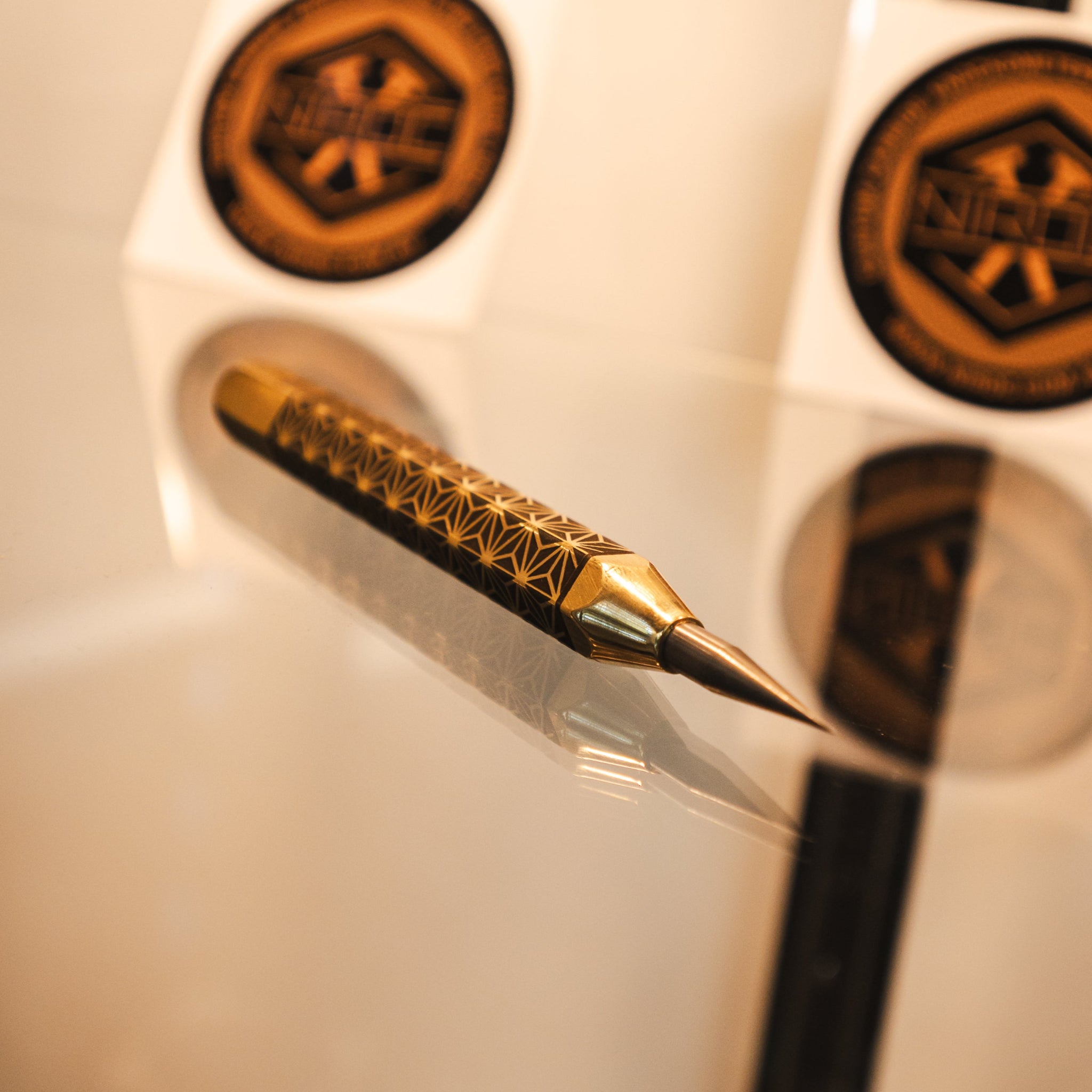 A gold and black decorative scribe with intricate geometric patterns on the anti-roll body, resting on a reflective glass surface. made out of brass and carbide.