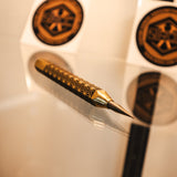 A gold and black decorative scribe with intricate geometric patterns on the anti-roll body, resting on a reflective glass surface. made out of brass and carbide.