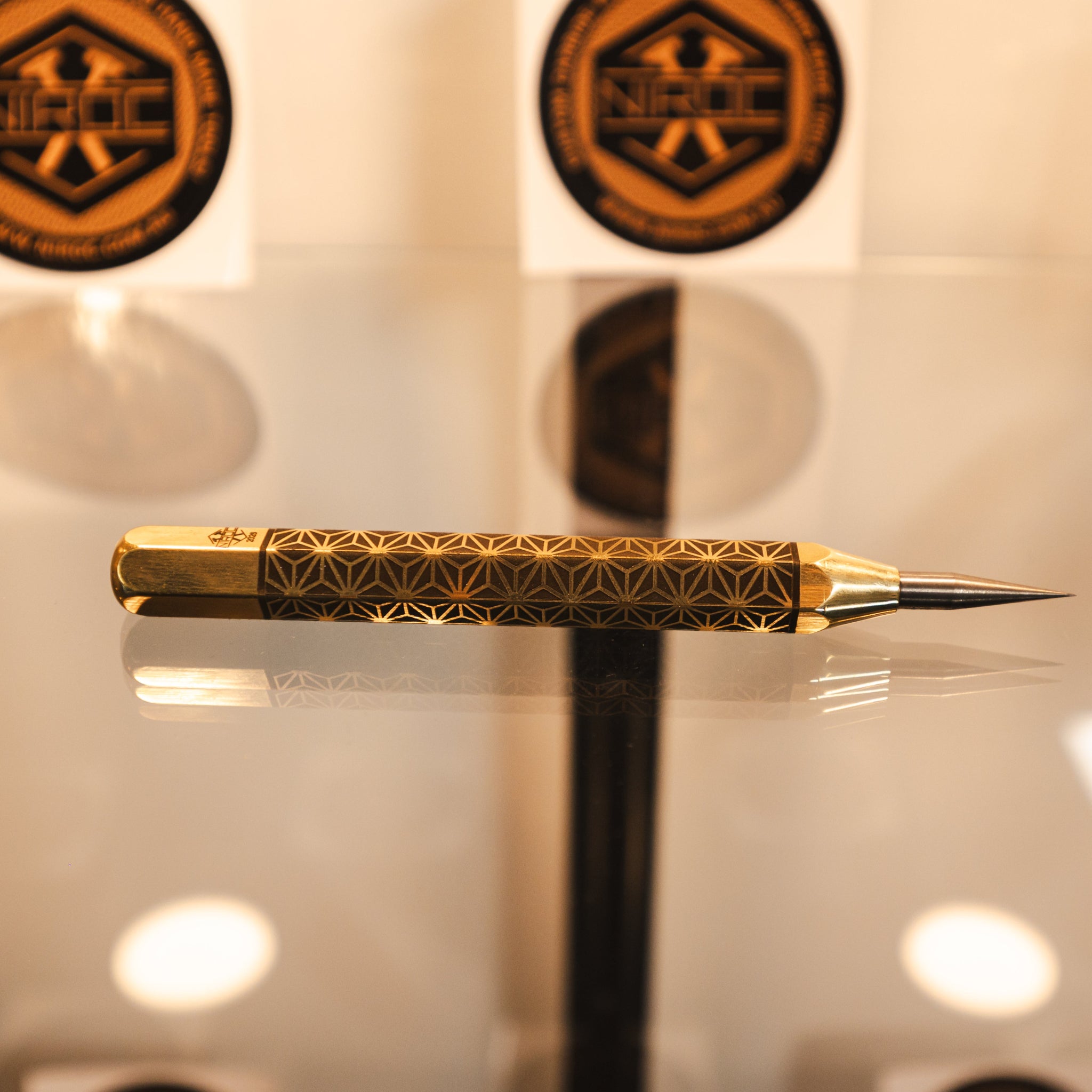 A gold and black patterned precision scribing tool, made out of brass and carbide resting on a reflective surface.