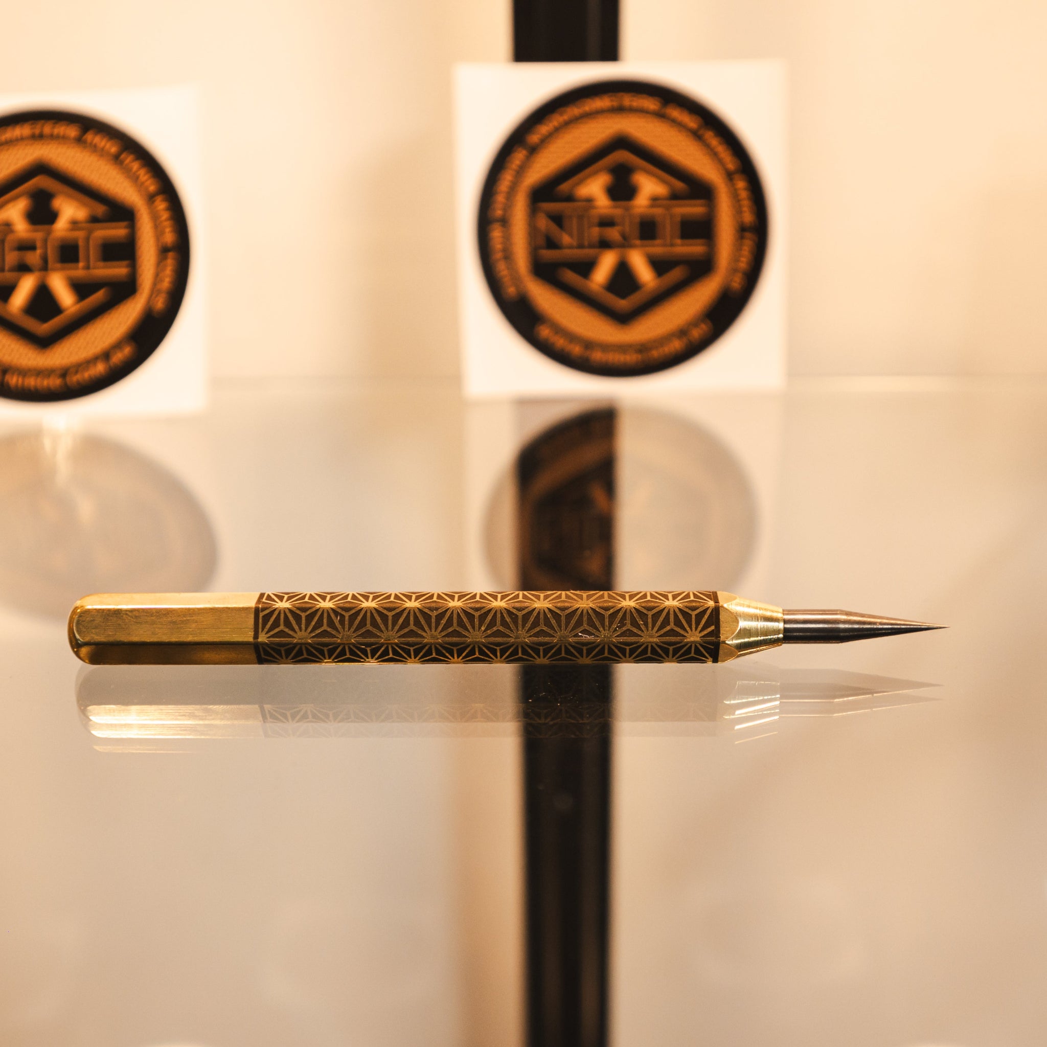 A gold-toned metal precision scribing tool, with a patterned brass handle and carbide tip, resting on a reflective surface.