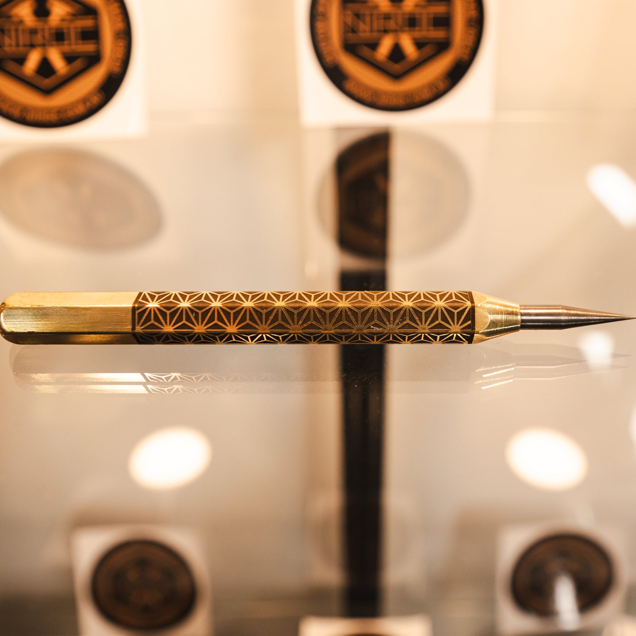 A brass scribe with intricate geometric pattern on its body and carbide tip, resting horizontally on a reflective glass surface.