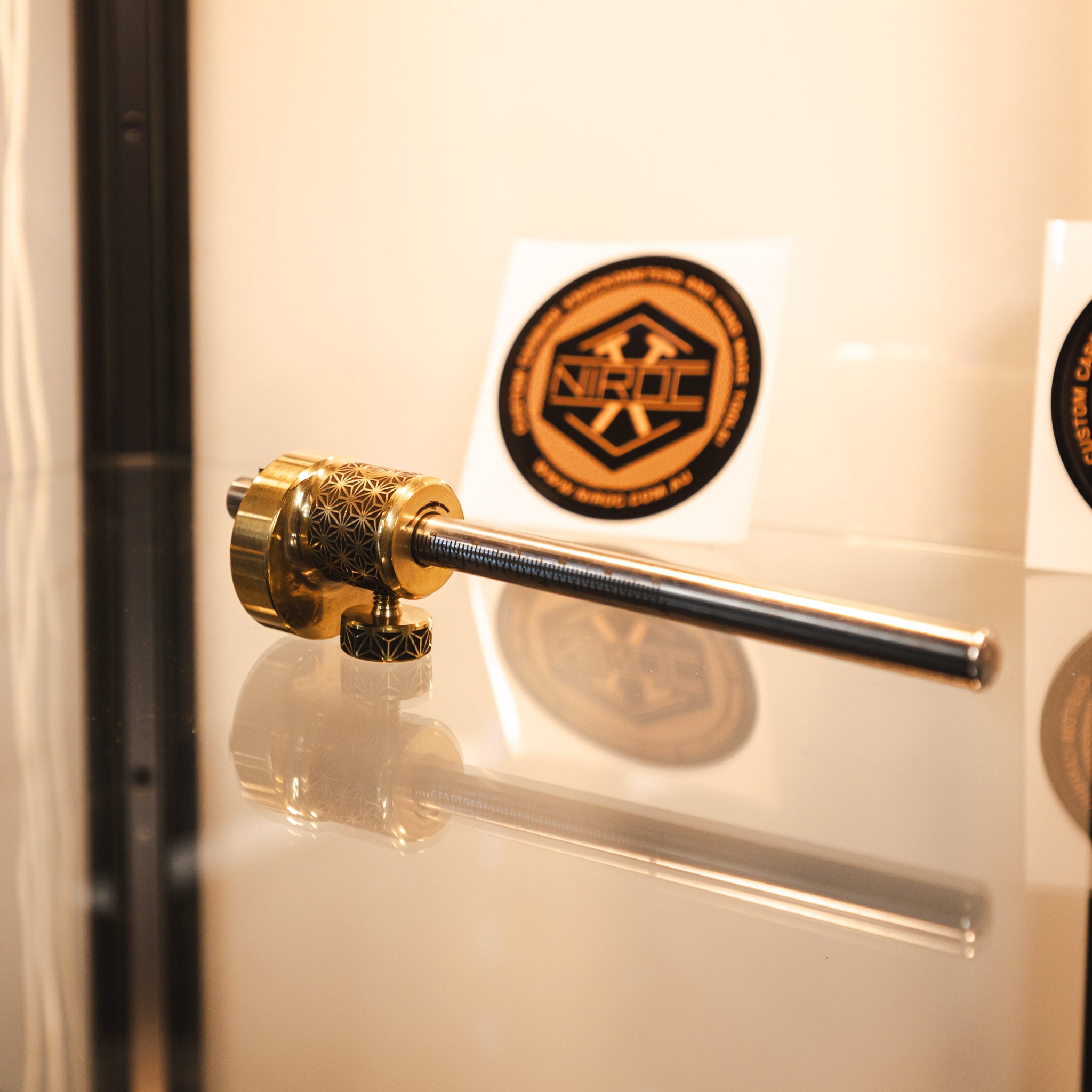 A brass and stainless steel marking gauge, displayed on a glass shelf with a warm beige background and circular stickers.