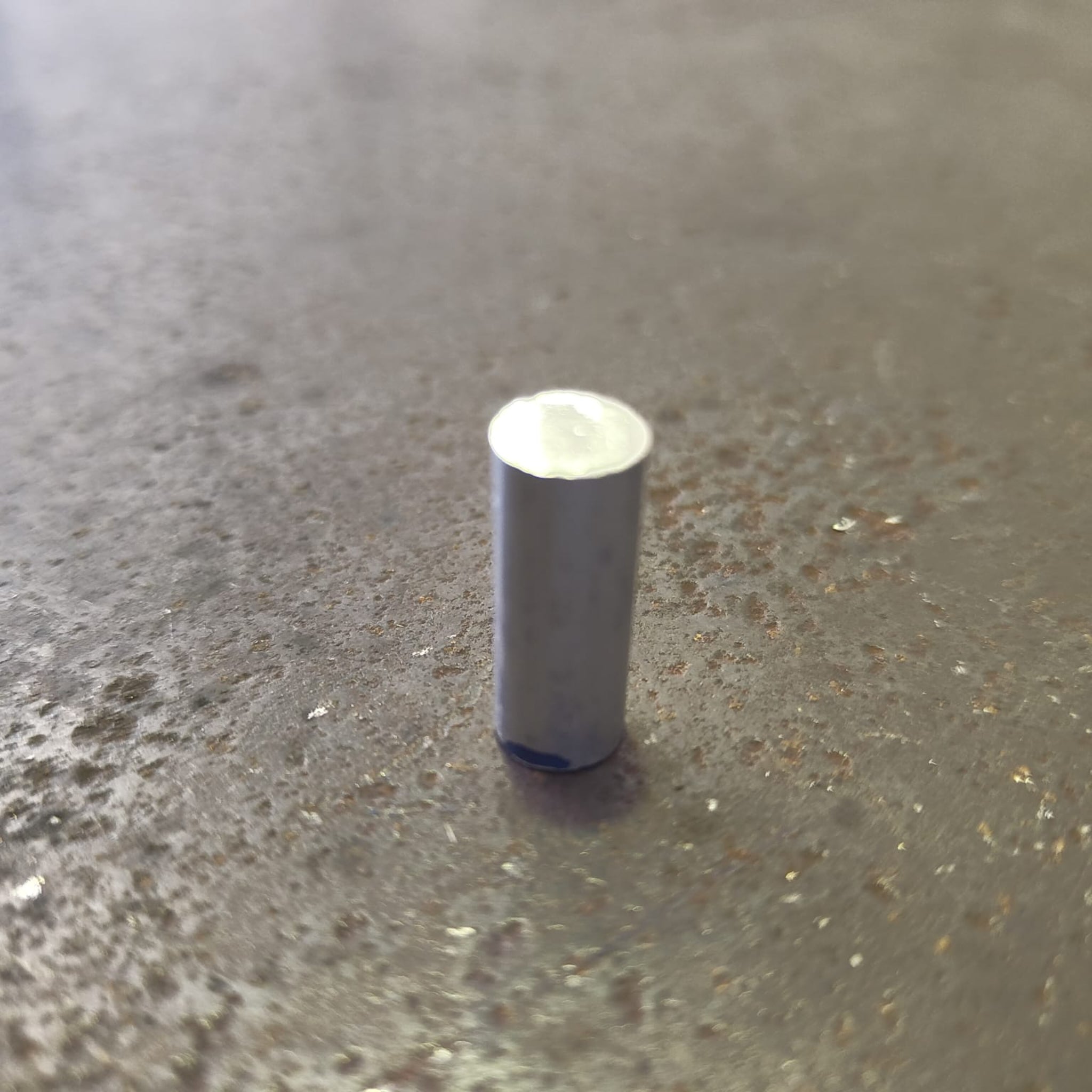 A small cylindrical metal object with a silver top and a dark gray body, standing upright on a textured steel bench.