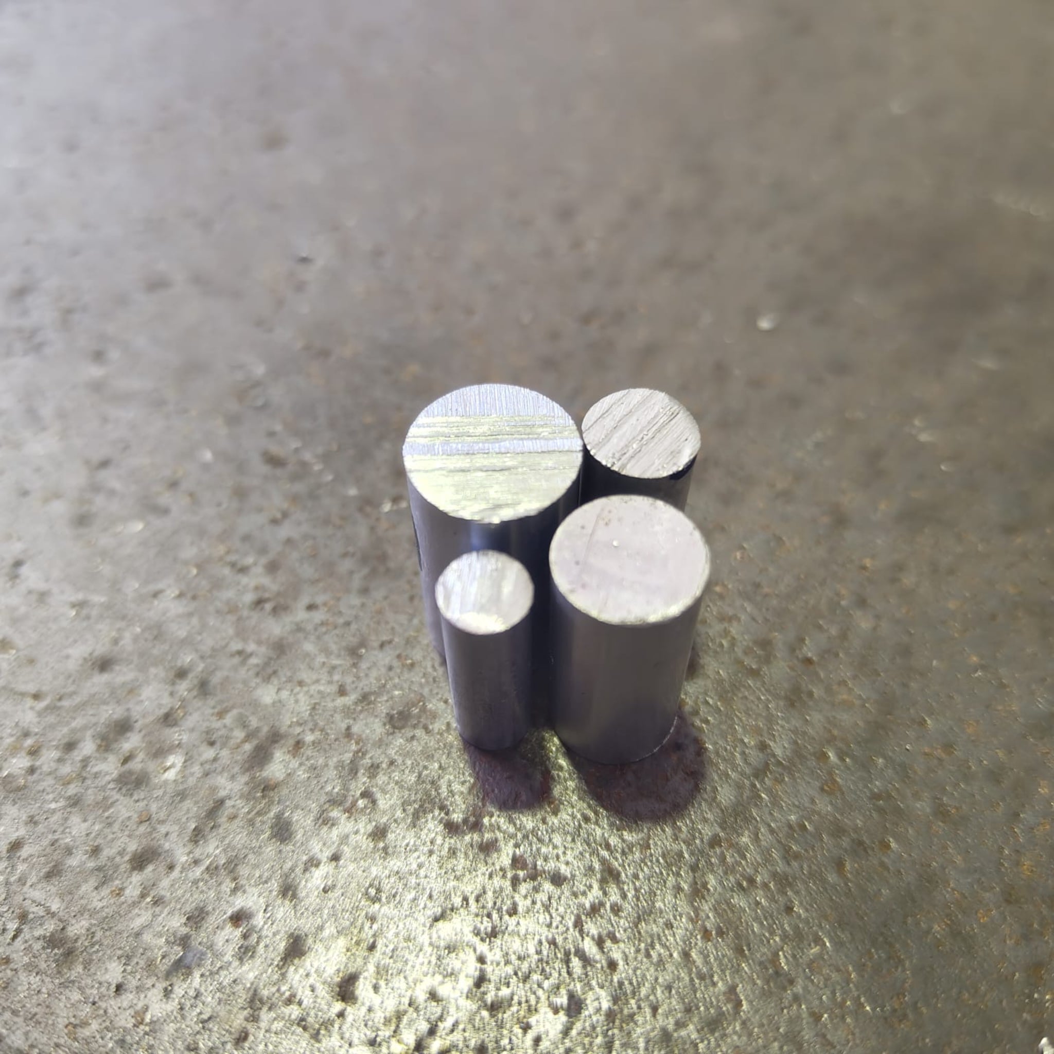 Four small cylindrical aluminium rods of different heights standing upright on a textured steel bench.