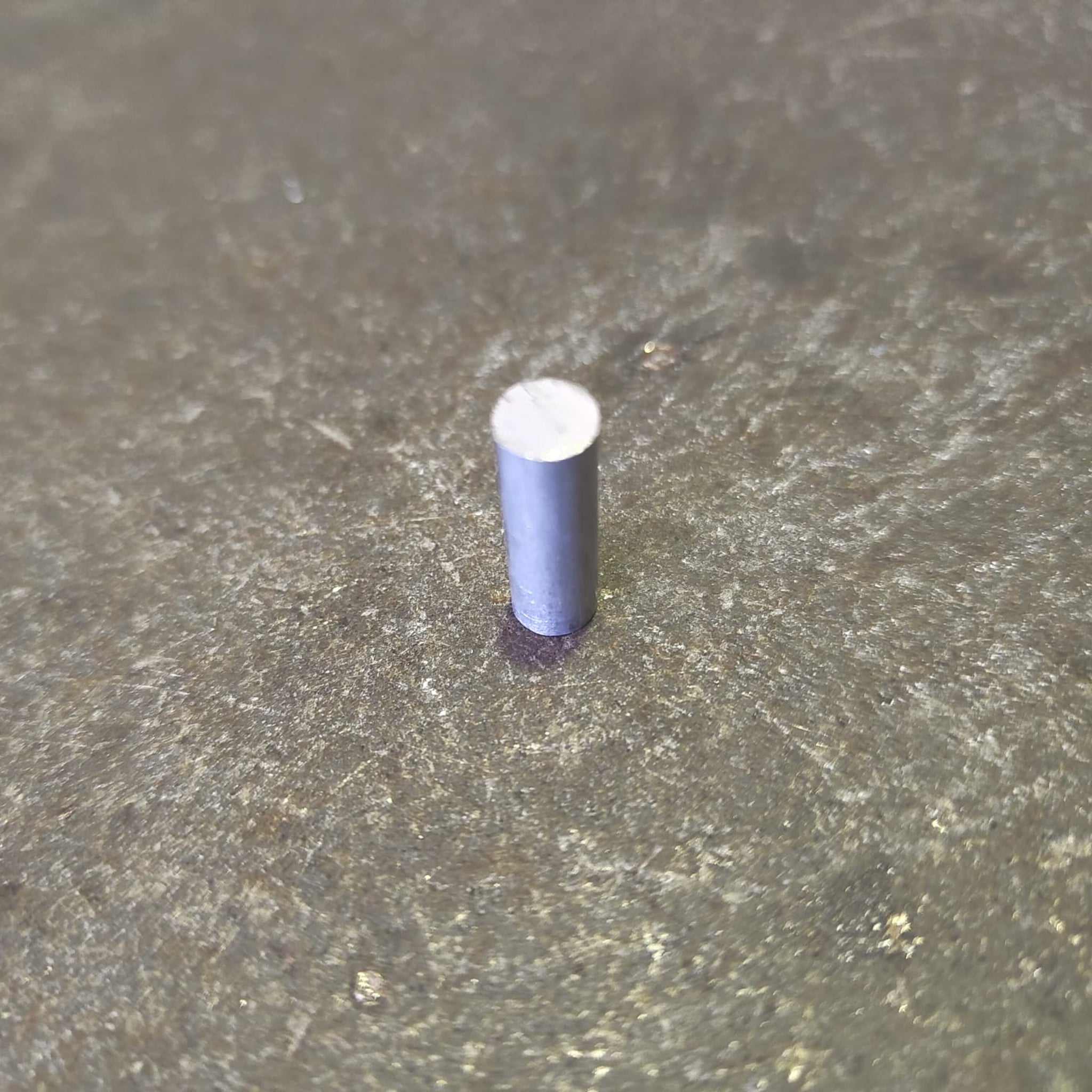 A small cylindrical aluminium rod or pin with a silver top, standing upright on a textured gray surface.