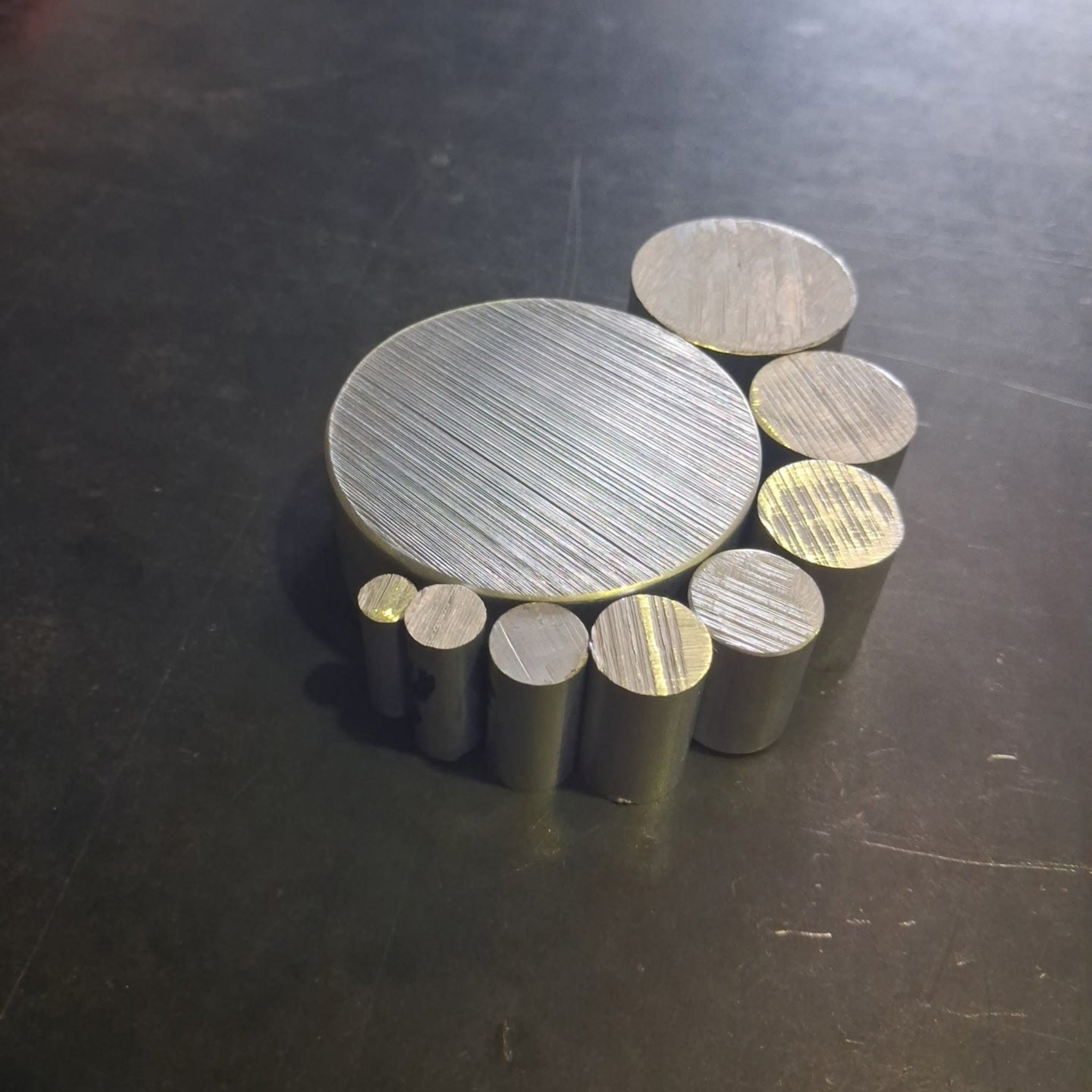 A set of aluminium cylindrical rods and discs in varying diameters, arranged on a dark, scratched workbench.