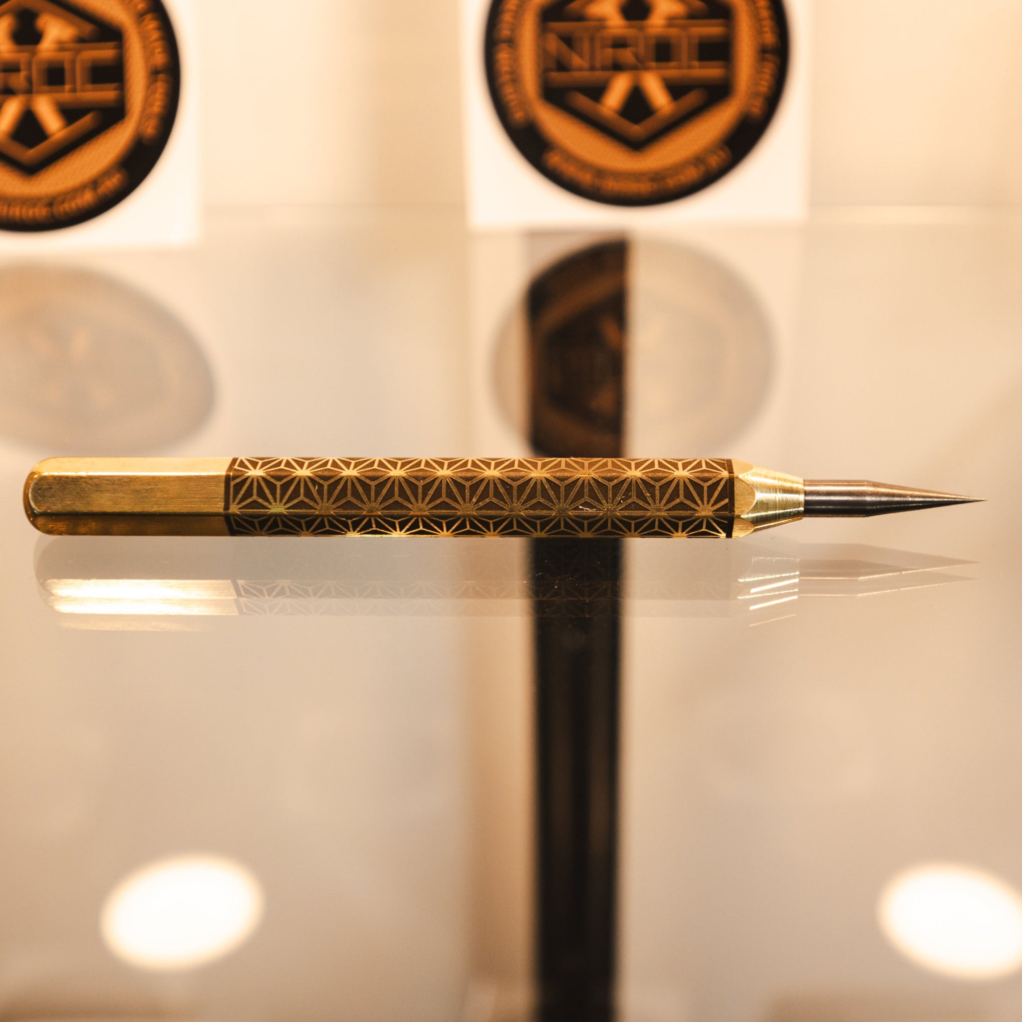 A gold-toned metal precision scribe tool, with an engraved handle and a fine carbide tip, resting on a reflective surface.