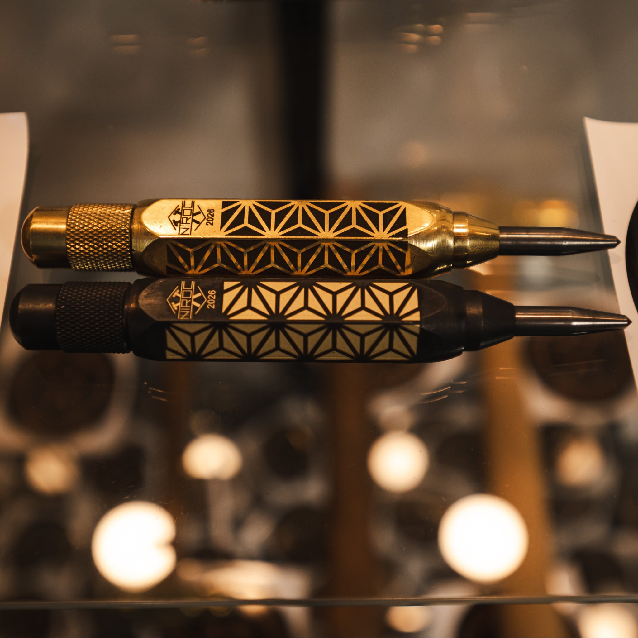 Two custom-engraved polished and black brass auto centre punches with geometric patterns, displayed horizontally on a glass surface with reflections