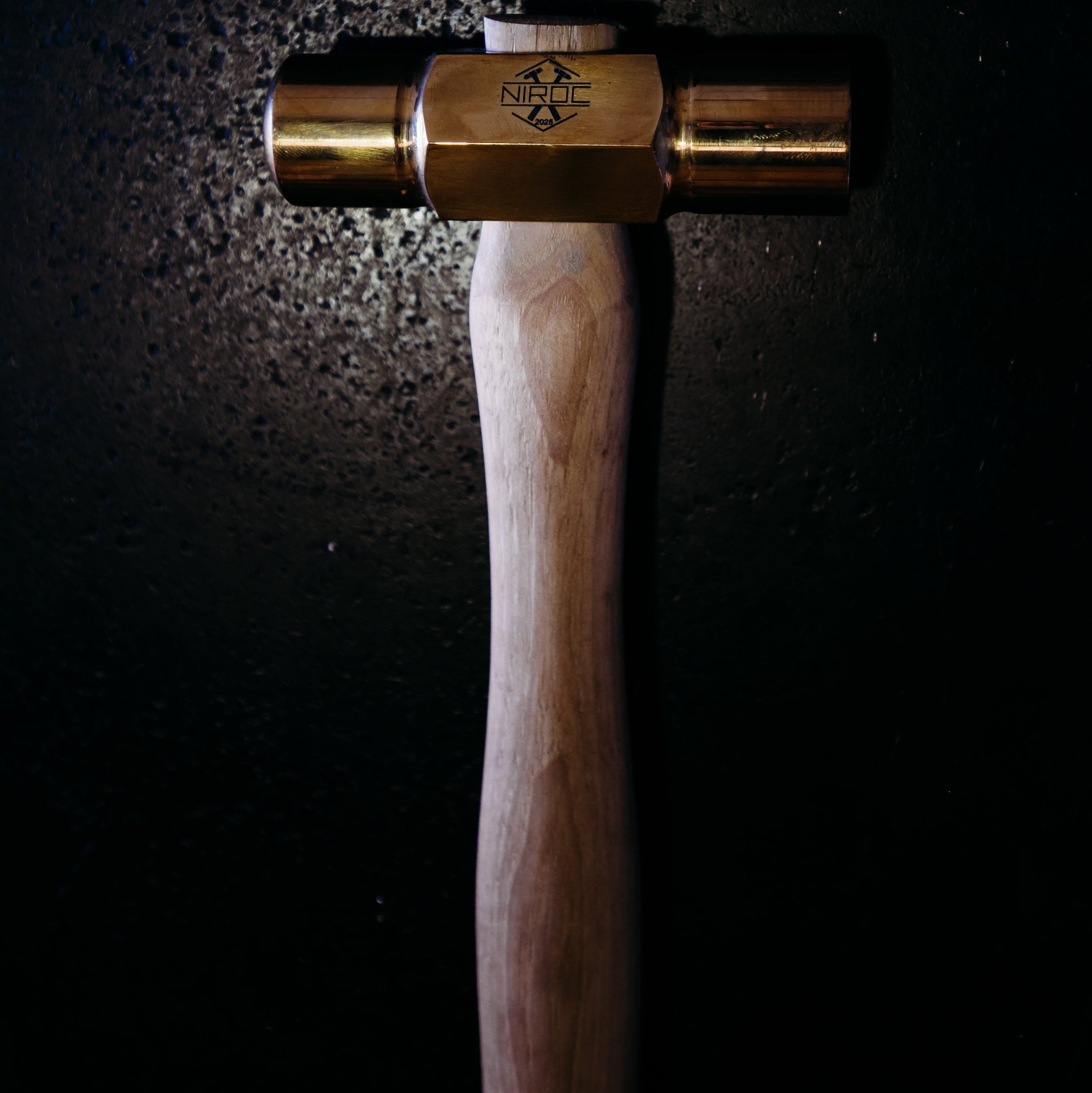 A brass and wood hammer photographed against a dark, textured background with dramatic lighting highlighting the metal head.