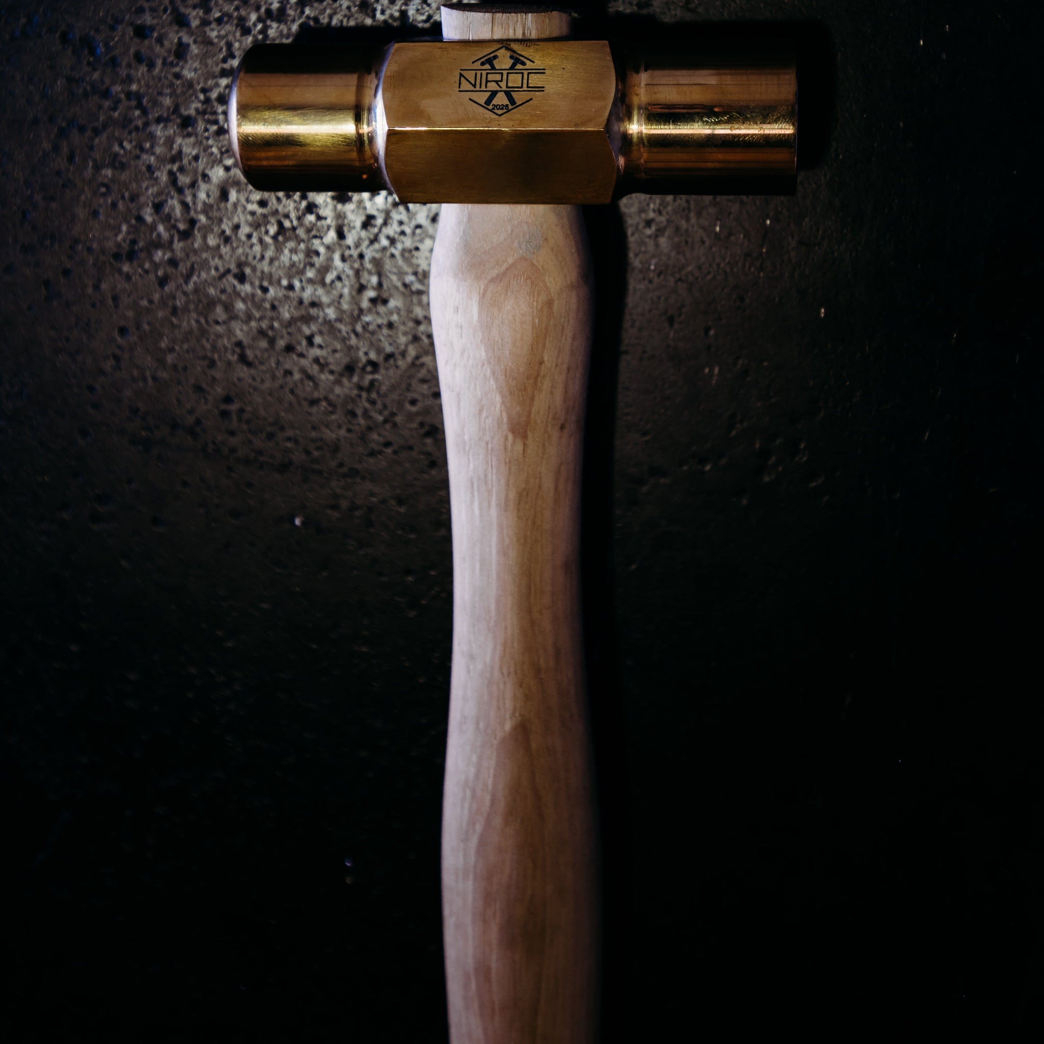 A close-up of a hammer with a brass head and a light wood handle, photographed against a dark, textured background with dramatic lighting.