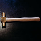 A brass hammer with a light-colored wooden handle, photographed horizontally on a dark, textured background with dramatic spotlighting.