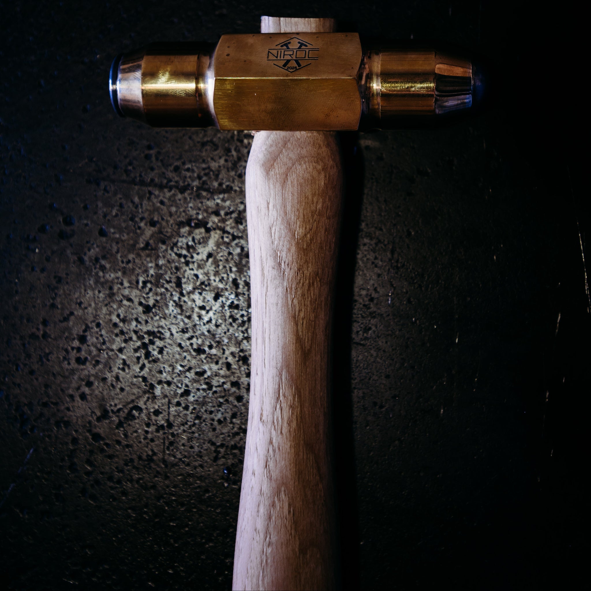 A close-up of a hammer with a brass-colored head and a light wood handle, positioned vertically against a dark, textured background.