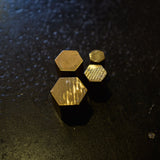 Four small hexagonal brass pieces in different sizes, arranged on a dark textured surface under dramatic lighting.