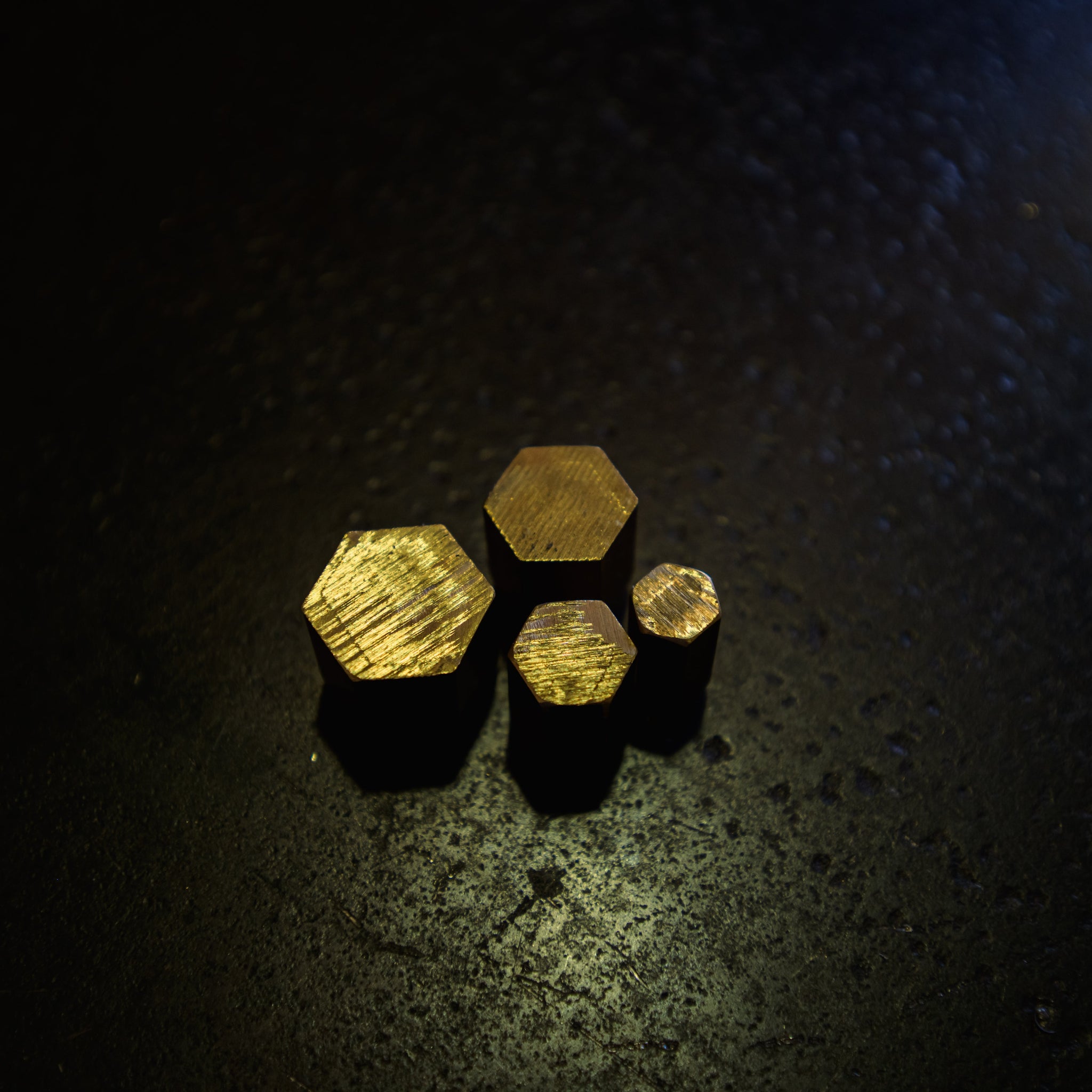 Four small hexagonal brass pieces with a brushed gold finish, arranged in a cluster on a dark, textured surface.
