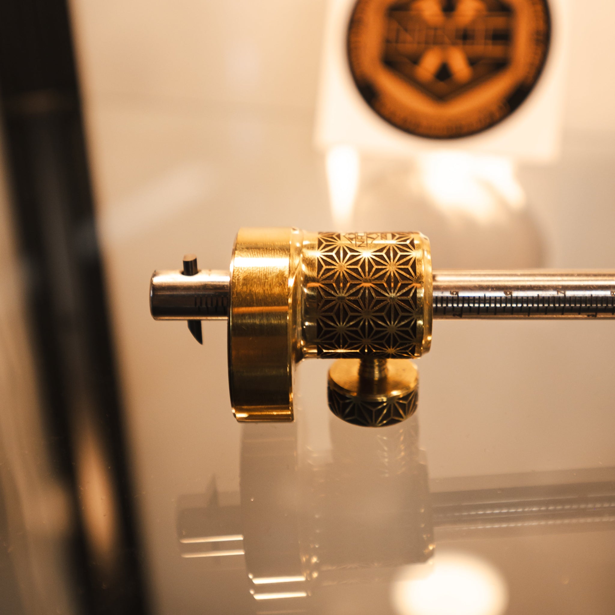 A close-up of a brass and stainless steel marking gauge with carbide tip. The background is warm and softly blurred, featuring a circular logo sticker.