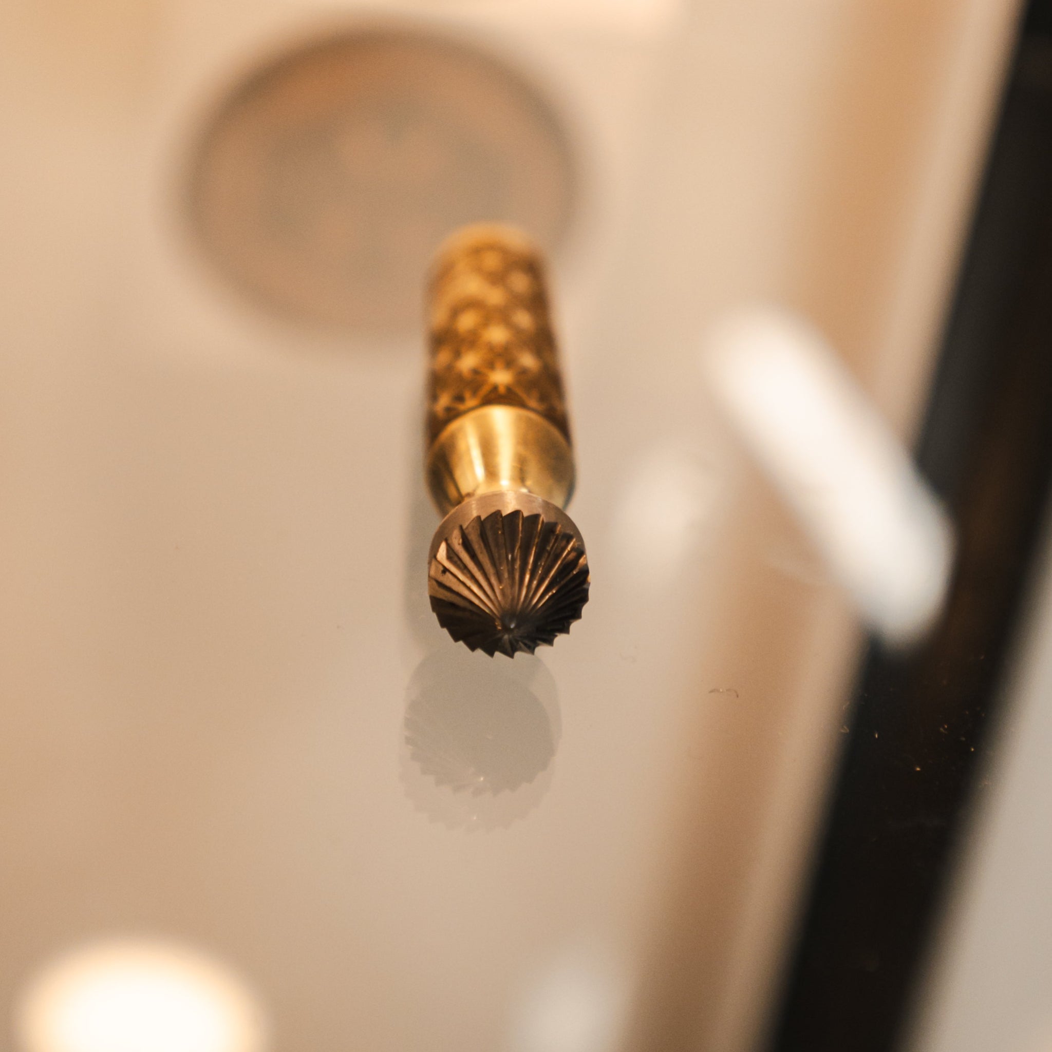 A close-up of a metallic gold and bronze textured cylindrical tool or accessory with a fluted dark tip, shown against a blurred light beige background.