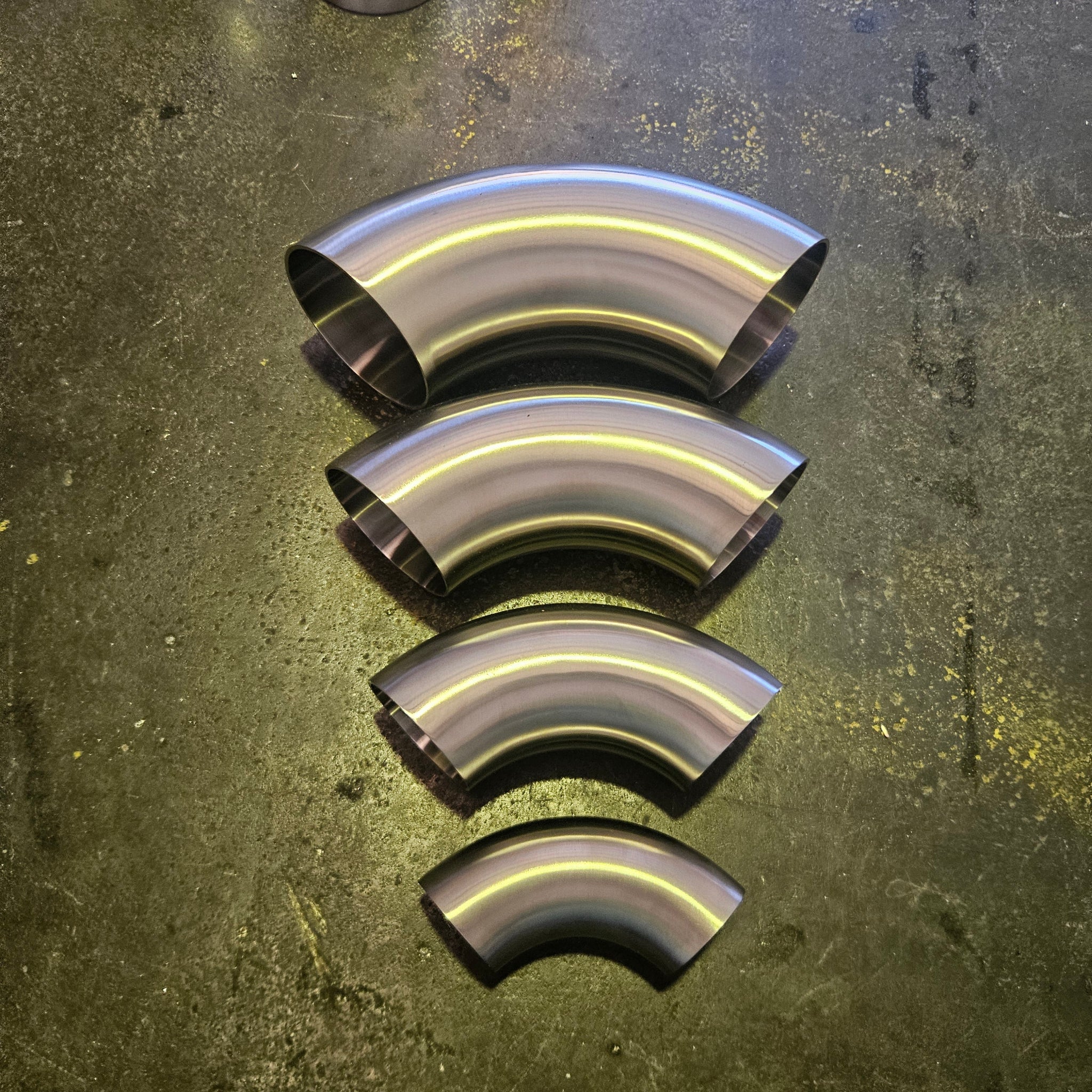 Four metallic curved pipe sections of different diameters arranged vertically on a dark, textured steel surface.