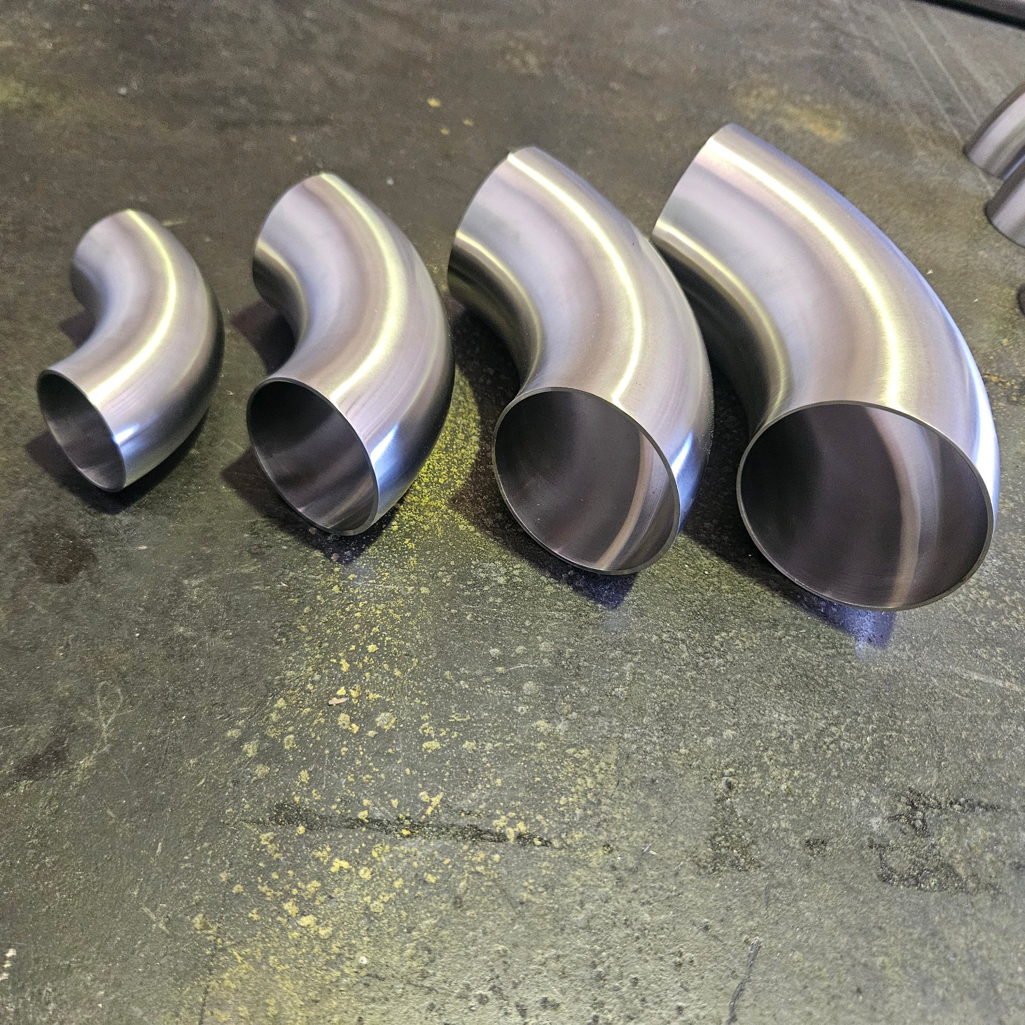 Four stainless steel 90 degree bends in different sizes arranged diagonally on a dark, textured workbench surface.