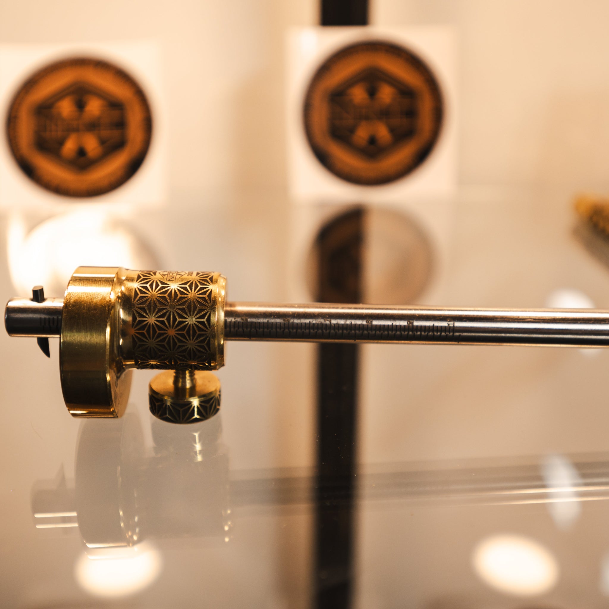 A brass and stainless steel marking gauge with a marked stainless rod and patterned brass collar, displayed on a reflective surface with blurred circular logos in the background.