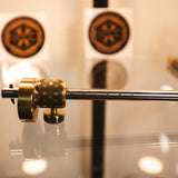 A brass and stainless steel marking gauge with a marked stainless rod and patterned brass collar, displayed on a reflective surface with blurred circular logos in the background.
