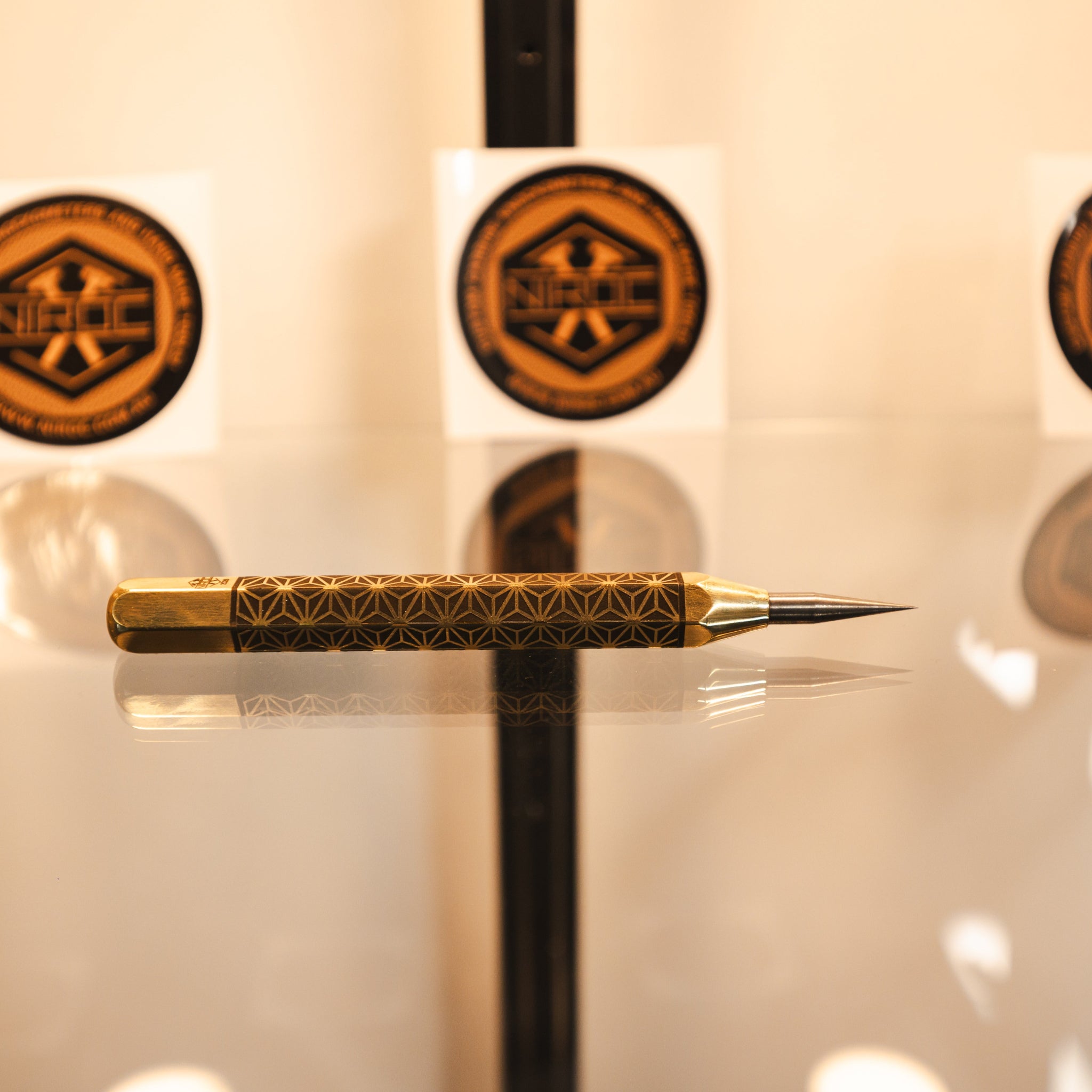 A gold and black patterned precision scribing tool, made out of brass and carbide, resting on a reflective surface.