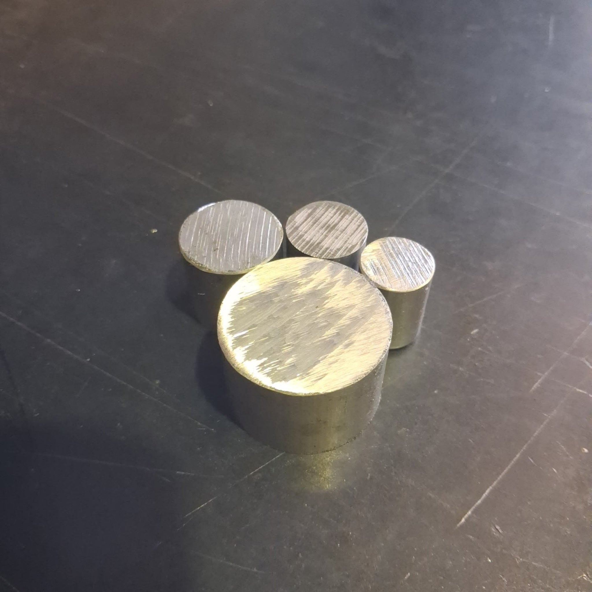 Four small cylindrical aluminium weights with textured tops arranged on a dark scratched surface.