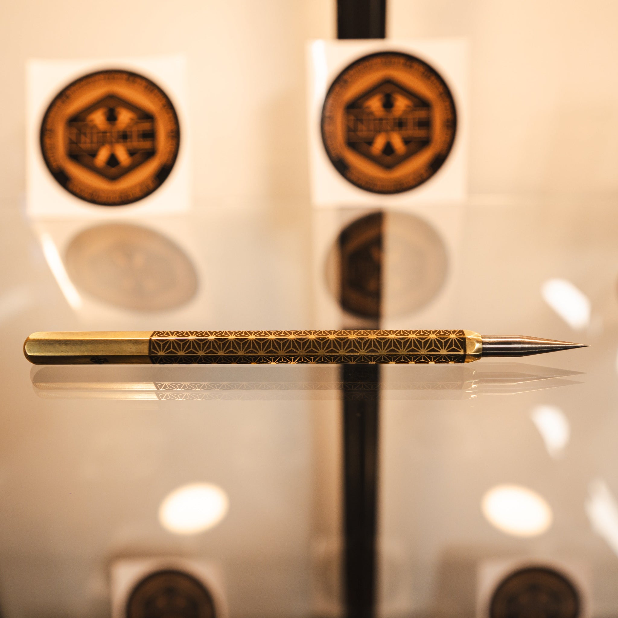 A gold and black patterned stylus pen resting horizontally on a reflective glass surface, with three matching circular gold and black emblem stickers displayed in the blurred background.