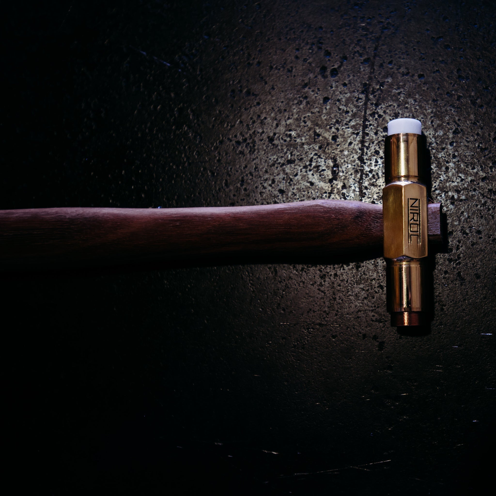 A brass soft face hammer with copper and nylon faces and a dark brown wooden handle, shown against a dark, textured background with dramatic lighting.