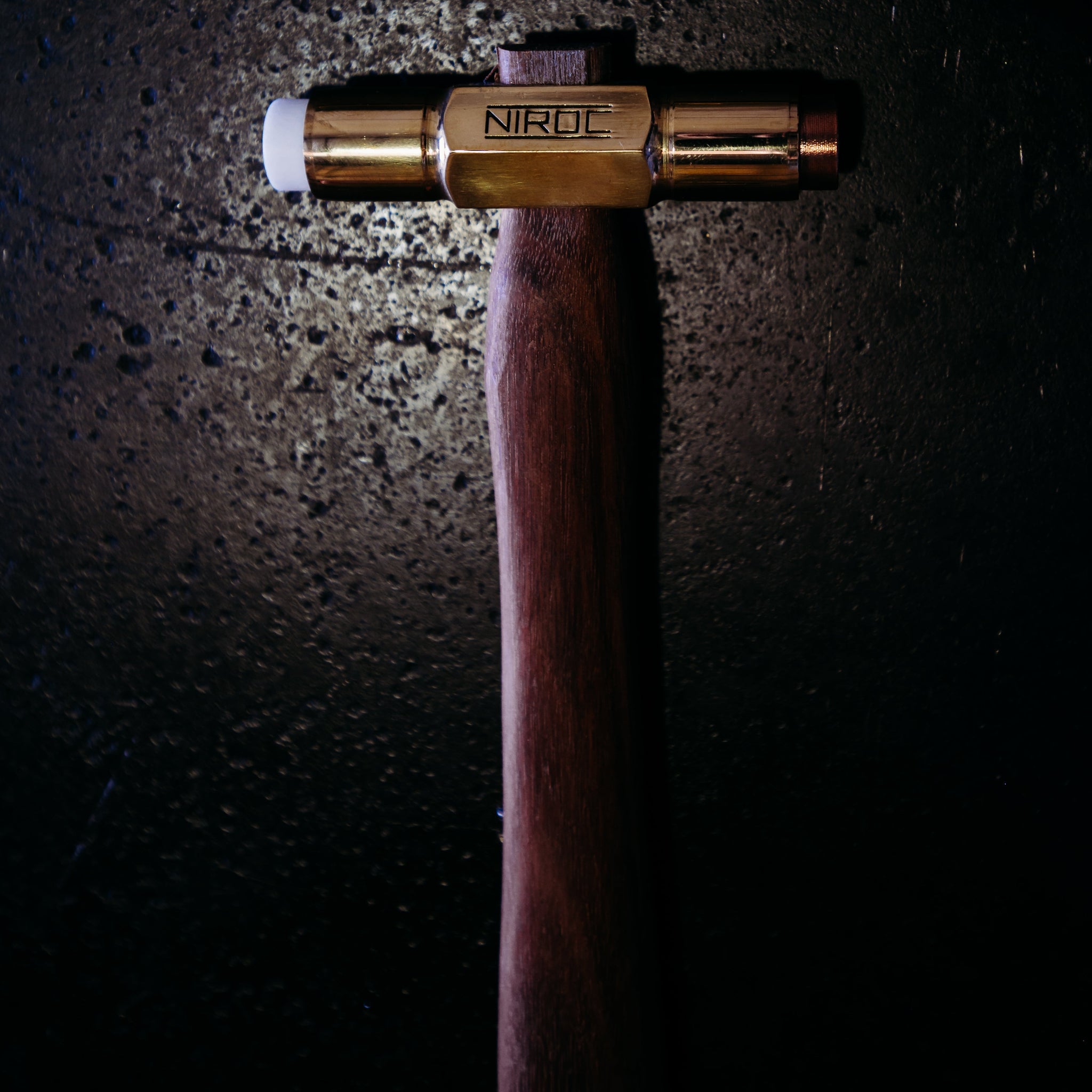 A close-up of a Niroc brand brass soft face hammer with a dark brown wooden handle, set against a dark, textured background with dramatic spotlighting.