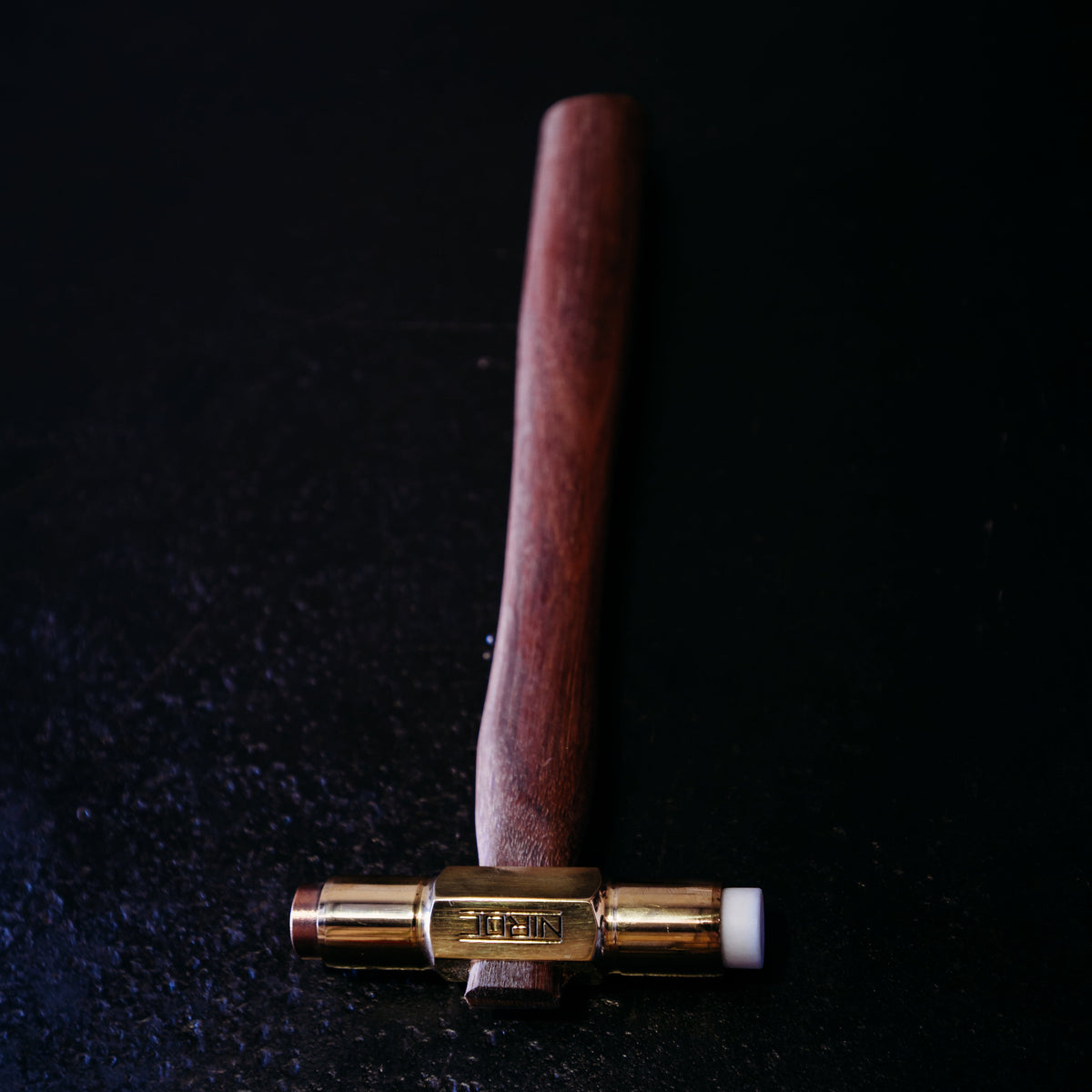 A wooden-handled hammer with a brass head and copper and nylon faces, photographed on a dark textured surface.