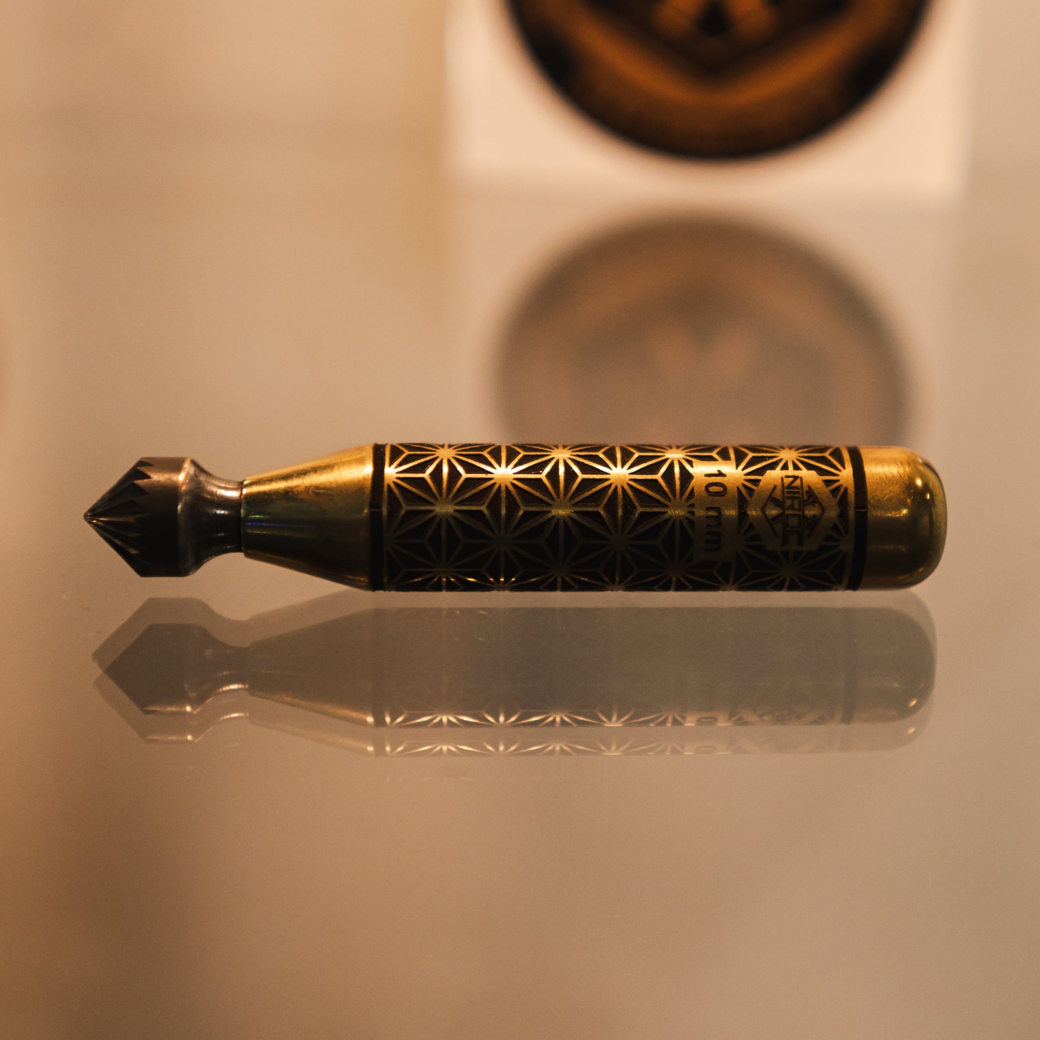 A decorative brass and black cylindrical tool or accessory with engraved geometric patterns, resting horizontally on a reflective surface with a blurred circular emblem in the background.