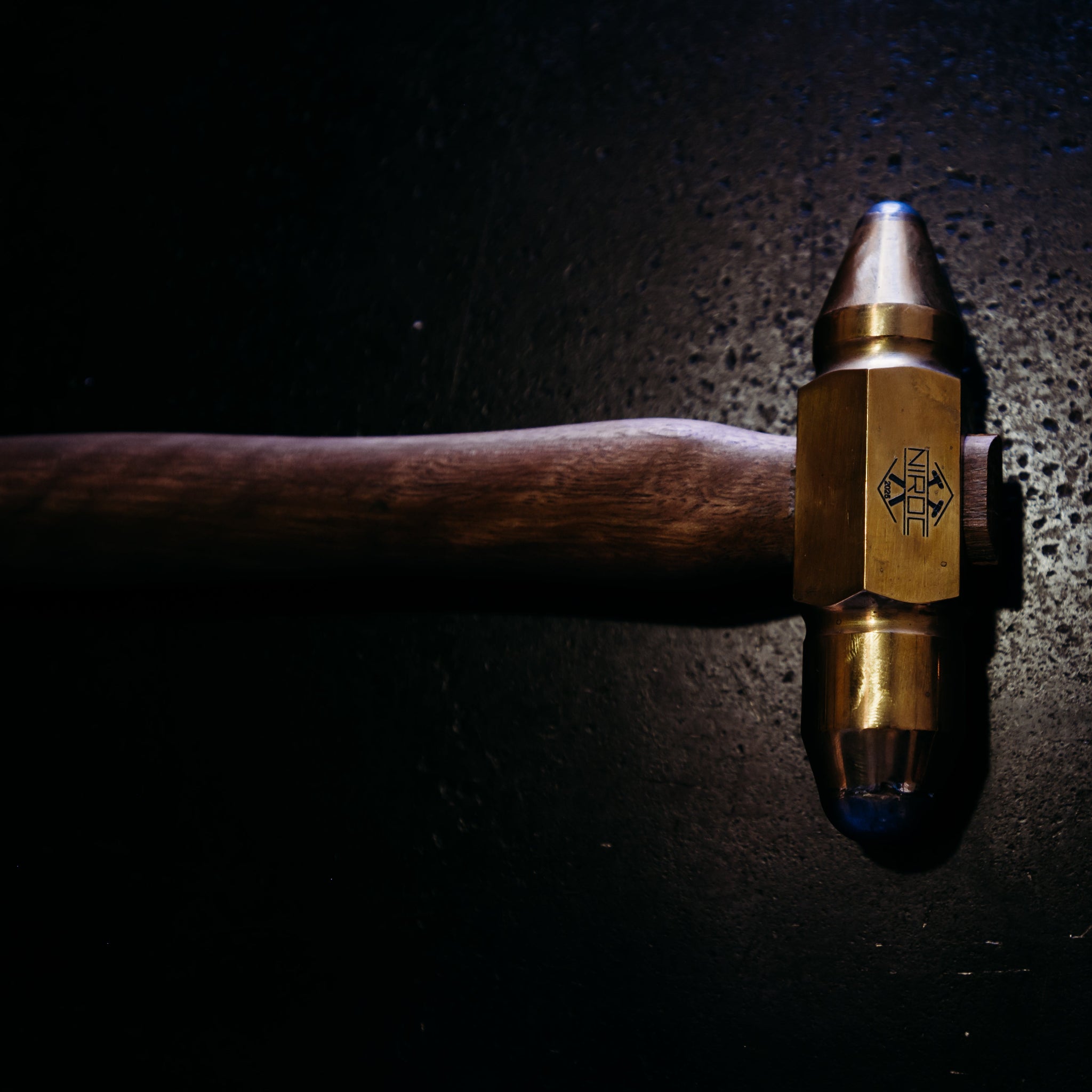 A close-up of a hammer head with a brass-colored hexagonal face and a dark wooden handle, set against a dark textured background with dramatic lighting.