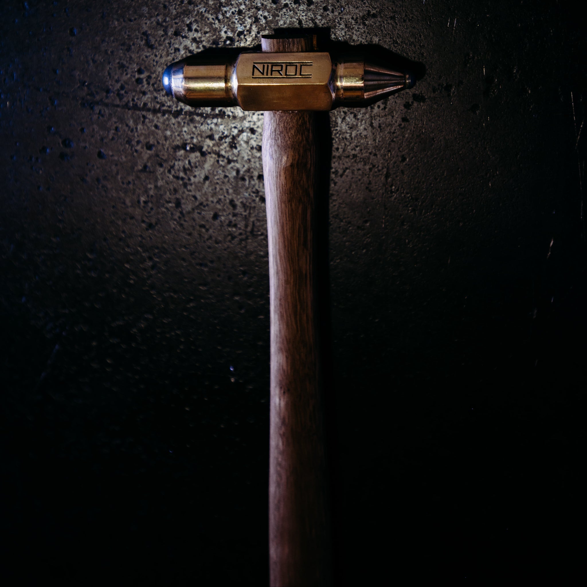 A custom metal hammer head made of brass and tungsten carbide , featuring a rounded tips and engraved with the brand name 'NIROC', mounted on a dark wooden handle against a dark, textured background.