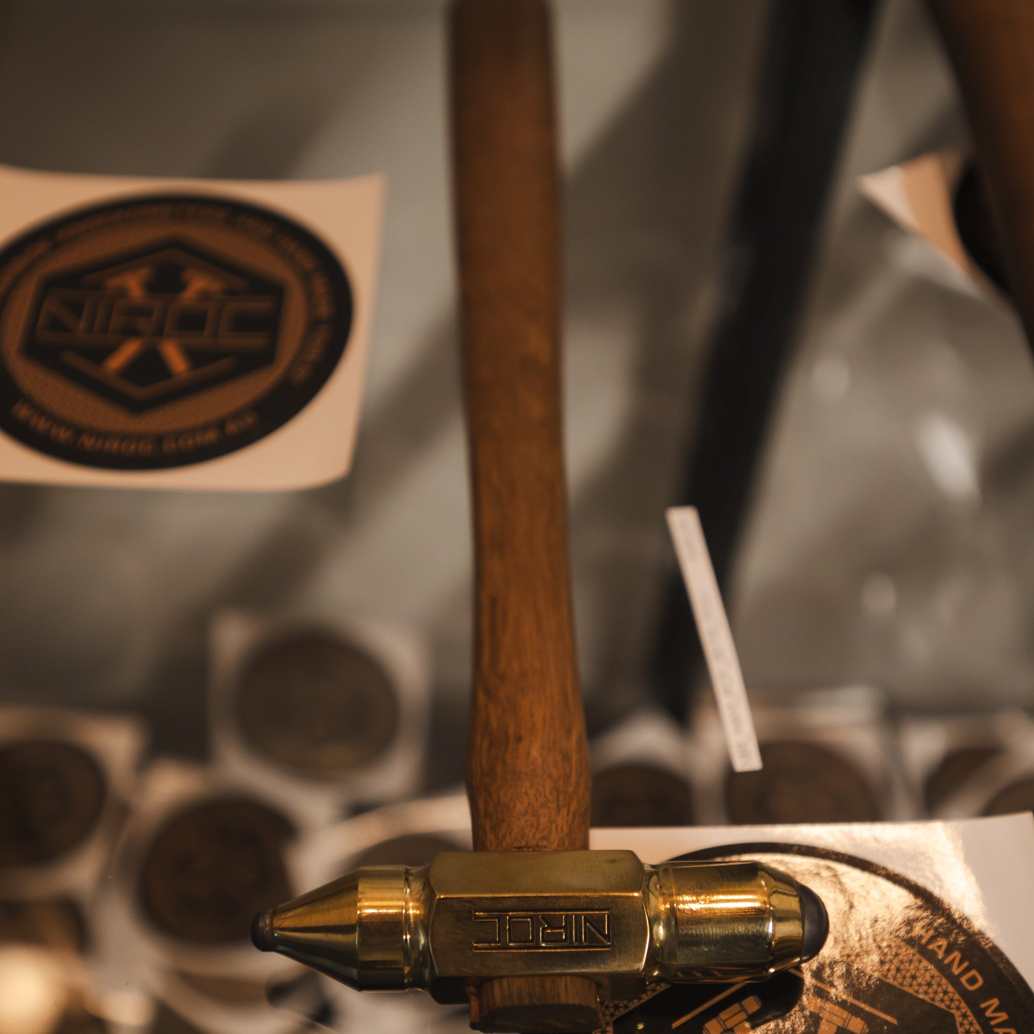 A close-up of a wooden-handled hammer with a brass head, resting on a circular metal display piece. The hammer head is engraved with the brand name 'NIROC'.