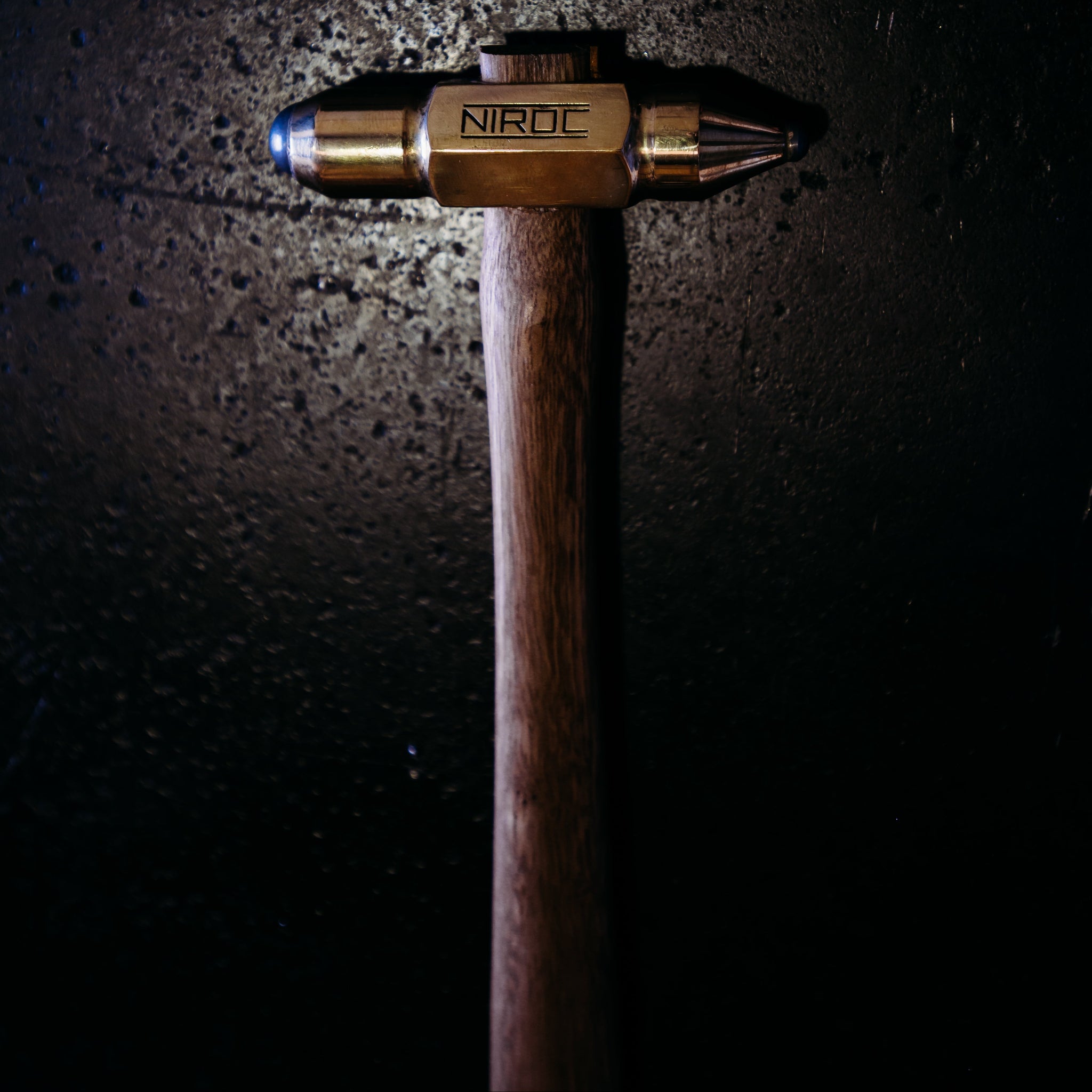A Niroc brand hammer with a polished brass head and a dark brown wooden handle, shown vertically against a dark, textured background with dramatic spotlighting.