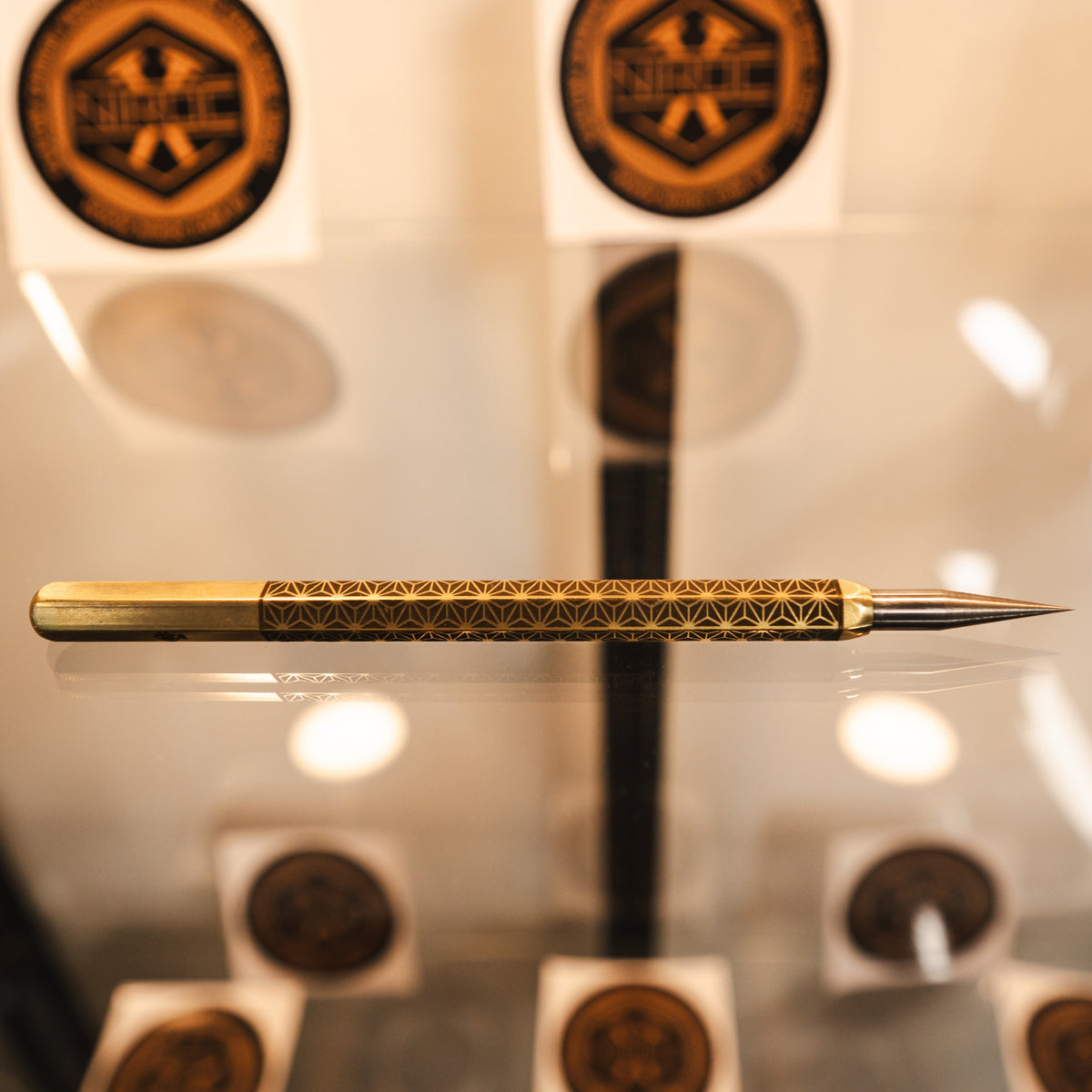 A polished brass scribe with intricate geometric engravings on its body and a sharp carbide tip, displayed horizontally on a reflective glass surface.