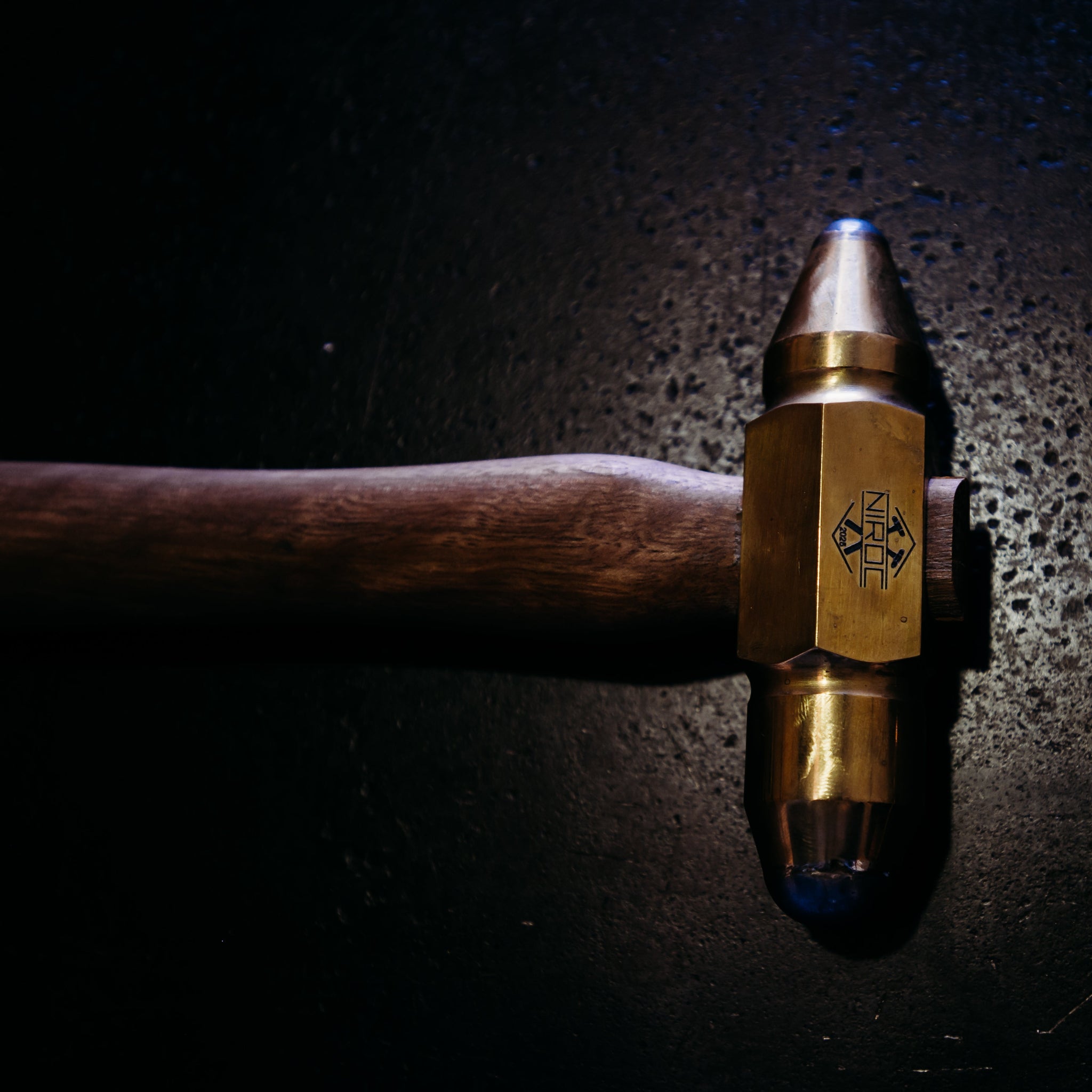 A close-up of a brass and wood hand tool, specialized tig hammer, resting on a dark textured background under dramatic spotlighting.